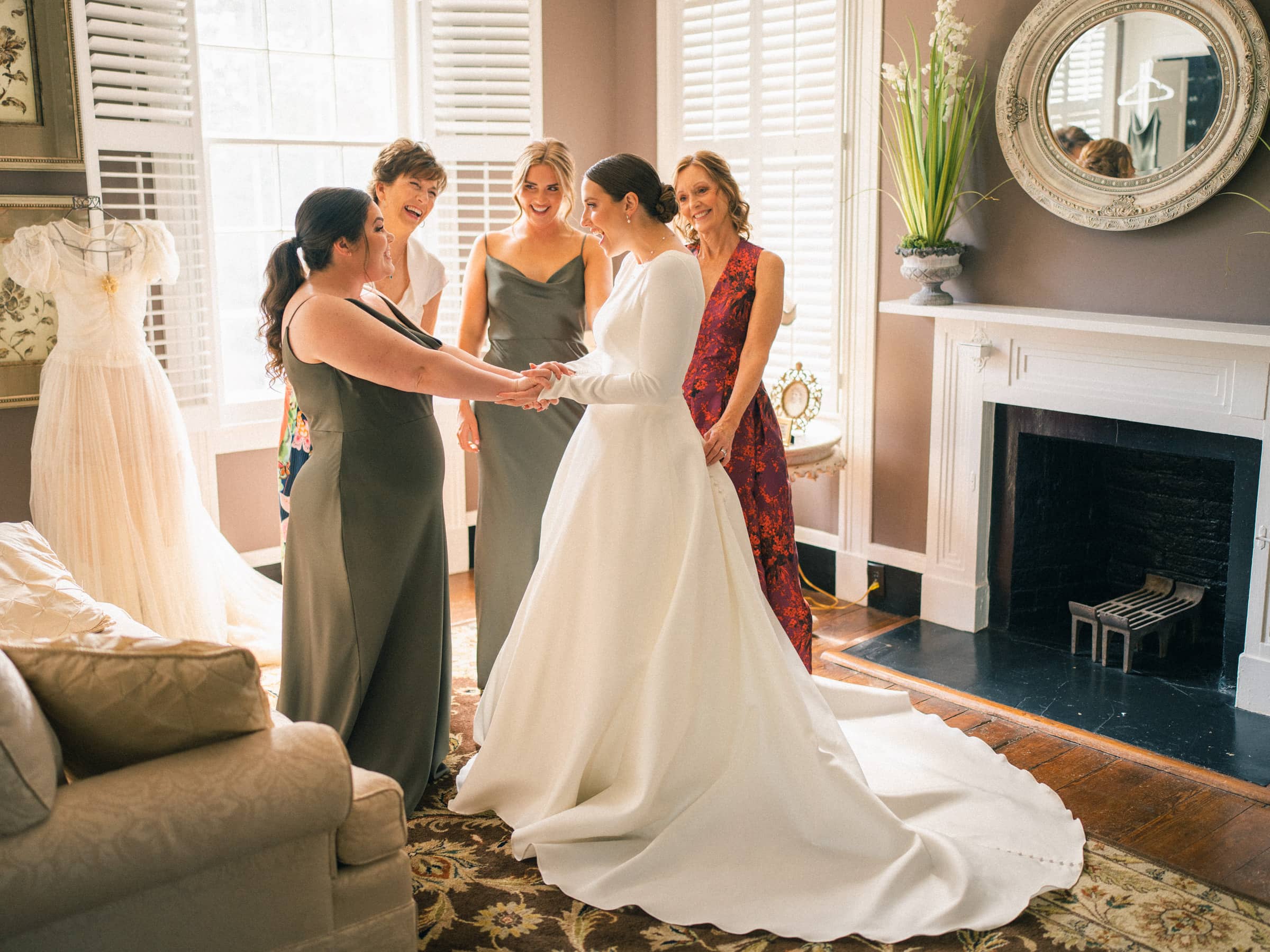 In a warmly lit room at Sutherland Estate, a bride in a white gown stands surrounded by four women, two in gray dresses and two in colorful ones. They smile as they admire her, while a mannequin adorned with a wedding dress lingers subtly in the background.