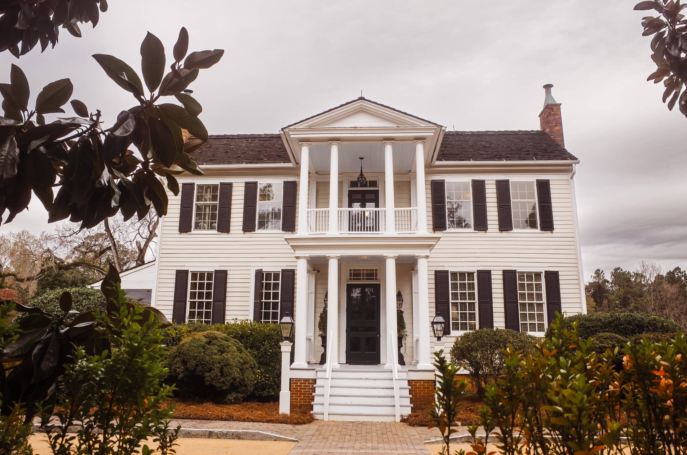 The Sutherland Estate, perfect for a wedding, features a large white colonial-style house with black shutters, a symmetrical facade, and a columned front porch. Surrounded by shrubs and trees under an overcast sky, it offers an elegant setting for your special day.