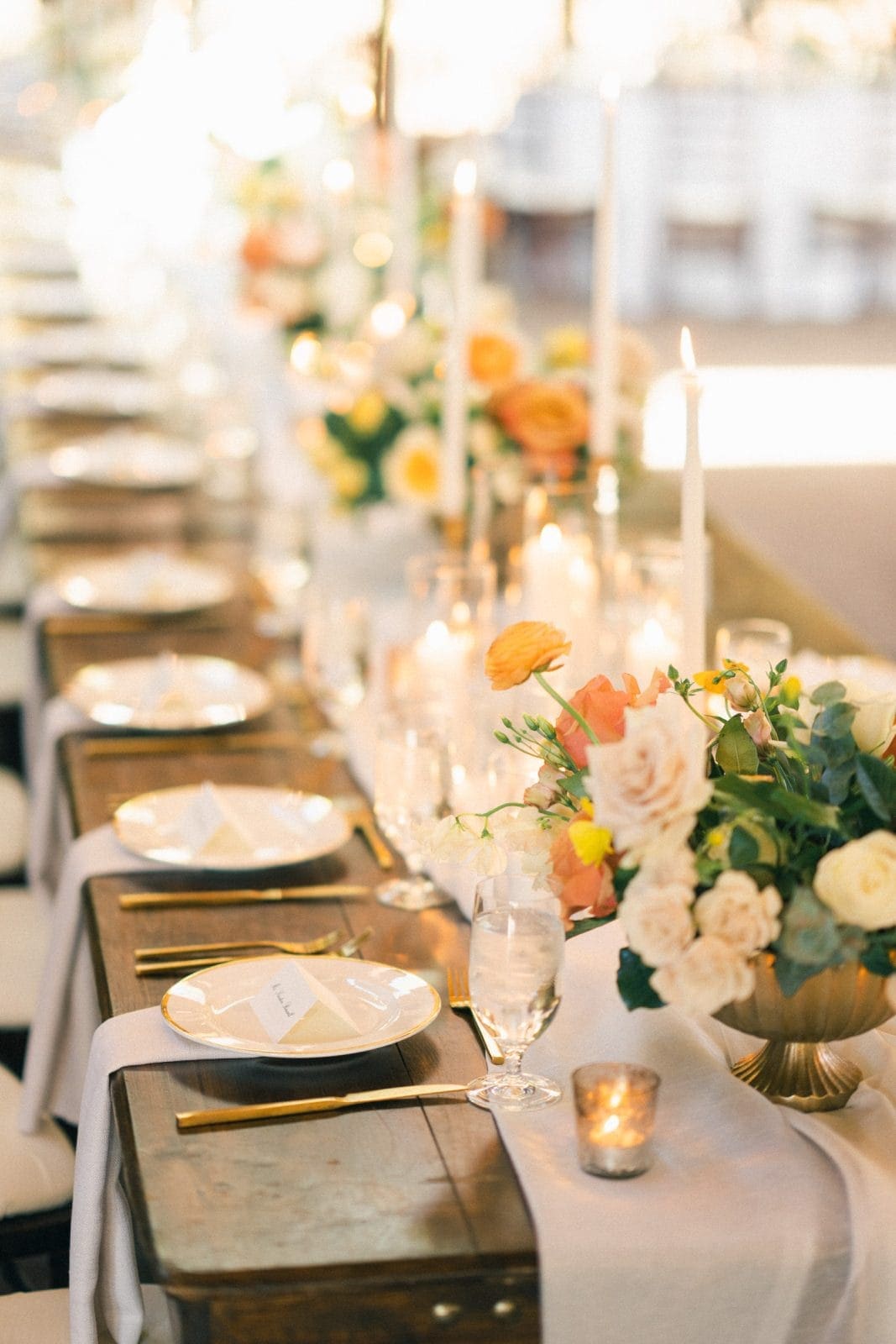 The elegantly decorated Sutherland Estate wedding reception table boasts white plates, gold cutlery, and crystal glasses. Centerpieces of orange and cream flowers, surrounded by glowing candles, create a warm ambiance perfectly suited for this enchanting venue.