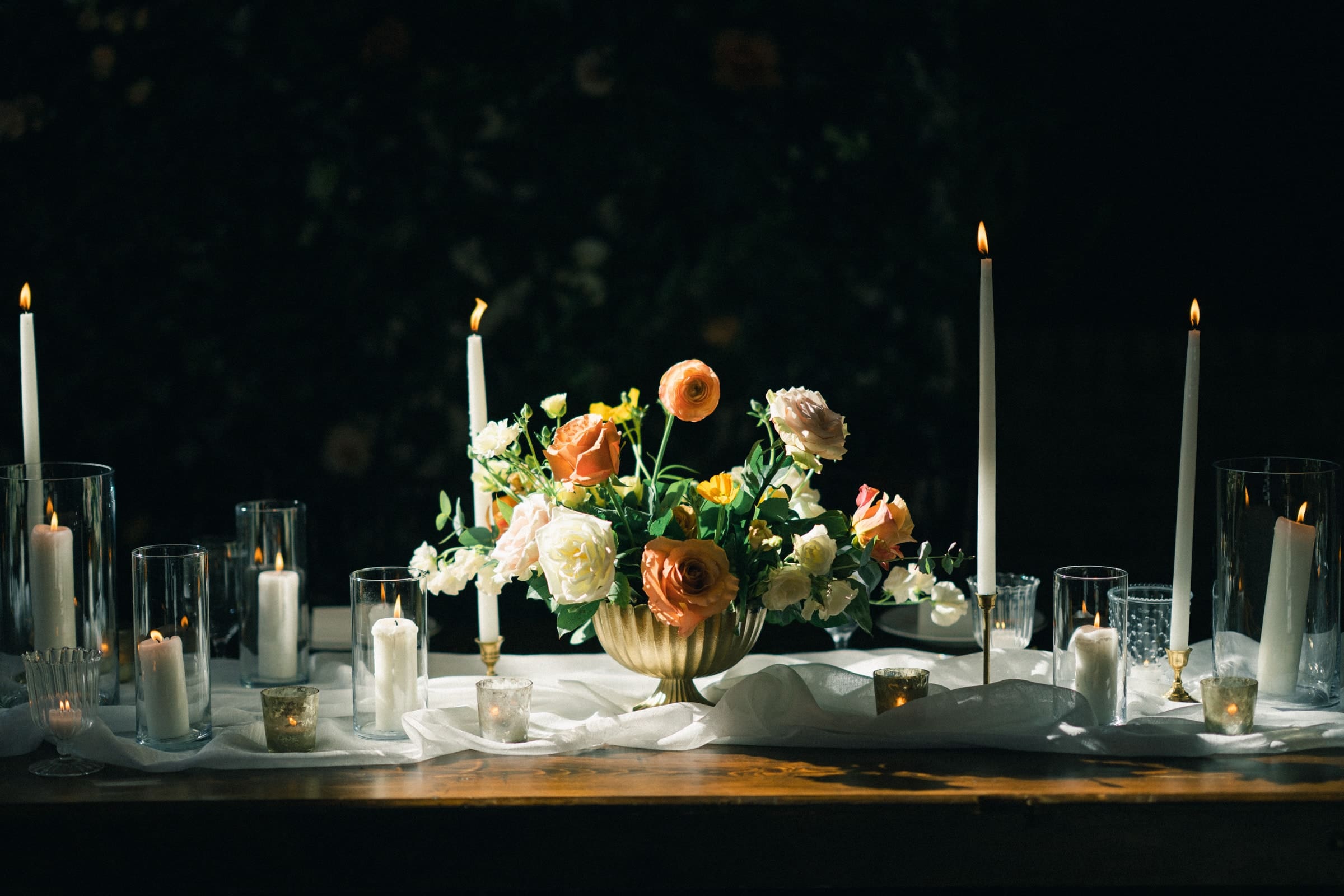 A dimly lit table at the Sutherland Estate wedding features a centerpiece of white and orange flowers. Tall and short candles in holders surround the arrangement, casting a warm glow. White fabric drapes softly across the dark wooden surface, creating an enchanting ambiance.