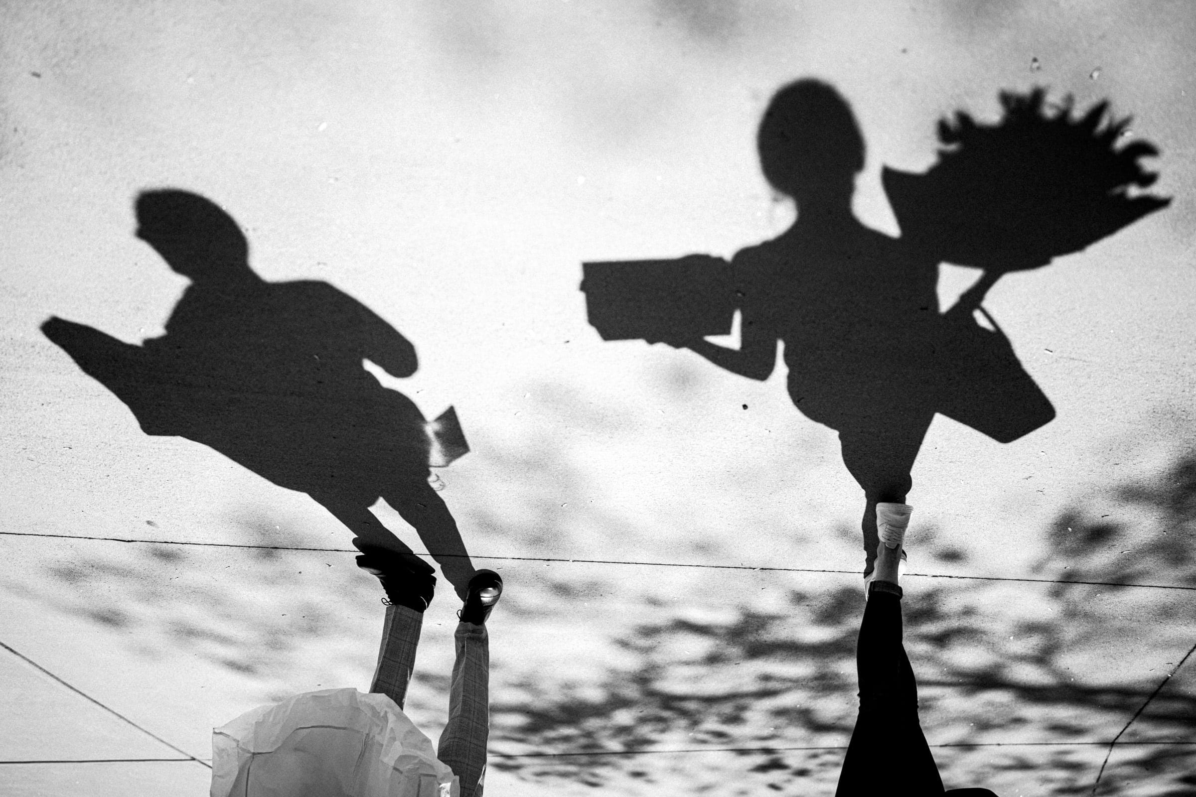 Shadows of a man and woman walking down a sidewalk hint at an unspoken story; the man clutches a book, while the woman holds a bouquet, reminiscent of an intimate elopement. Captured in black and white, blurred foliage shadows evoke the serenity of secluded meadows.