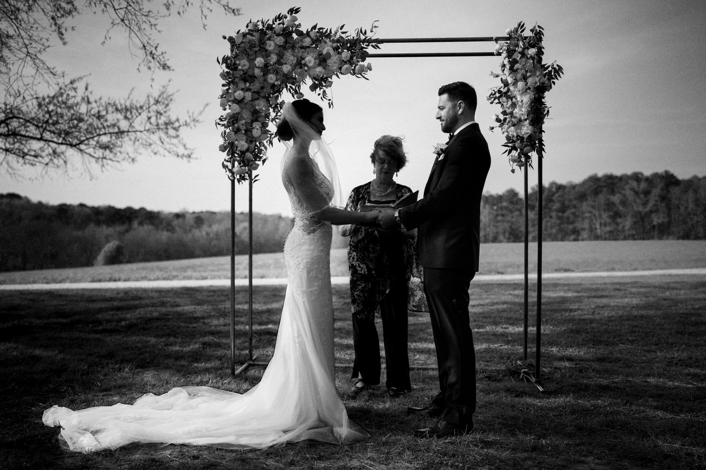 A bride and groom stand facing each other, holding hands under a floral arch in an intimate elopement. A woman stands between them, officiating the ceremony. The scene is serene, with a backdrop of trees and the sweeping beauty of the meadows beyond.