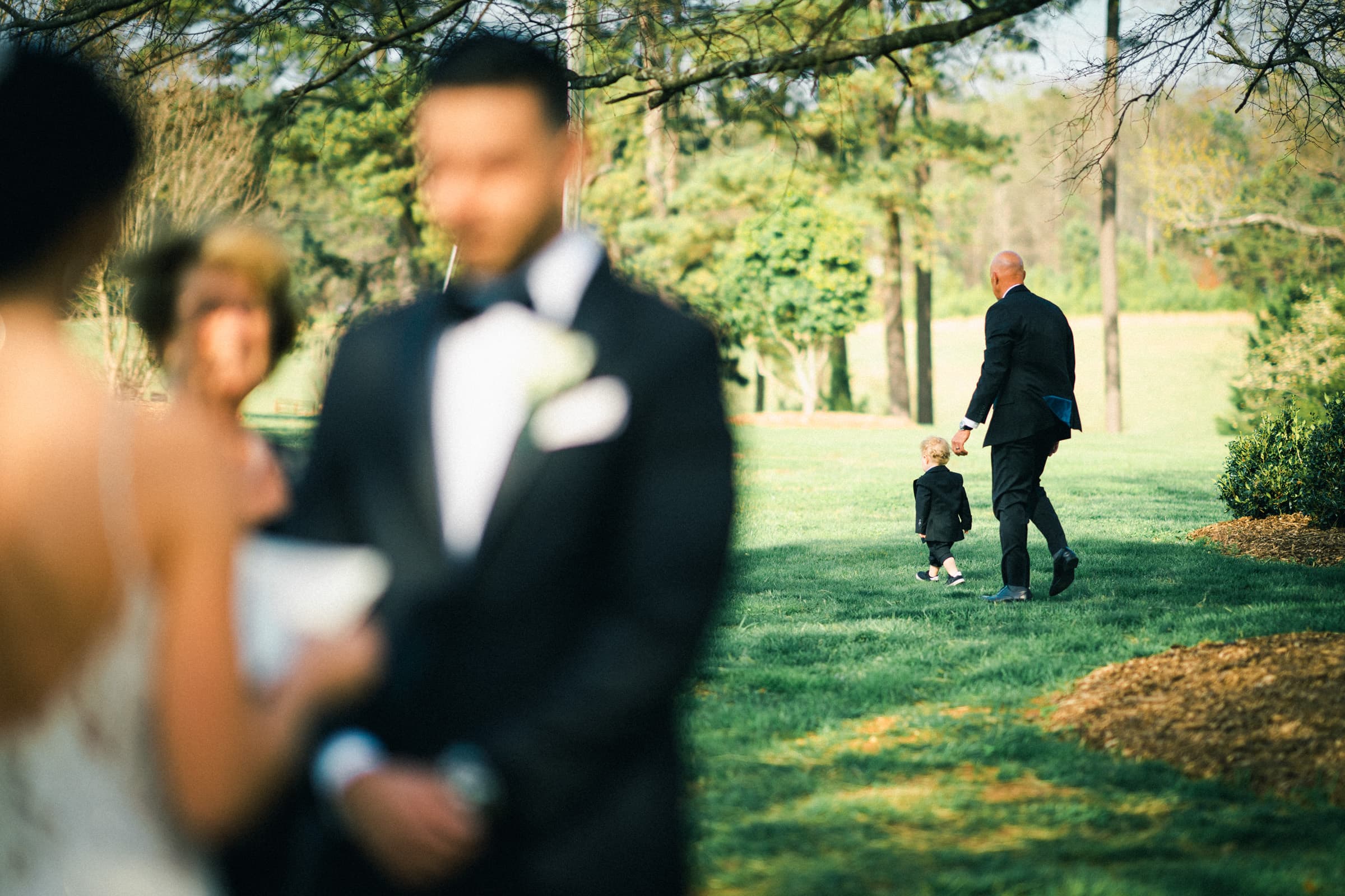 A wedding ceremony scene with a blurred bride and groom in the foreground captures the essence of an intimate meadows elopement. In the background, a man in a suit walks hand in hand with a toddler across a grassy area, surrounded by trees and soft sunlight.
