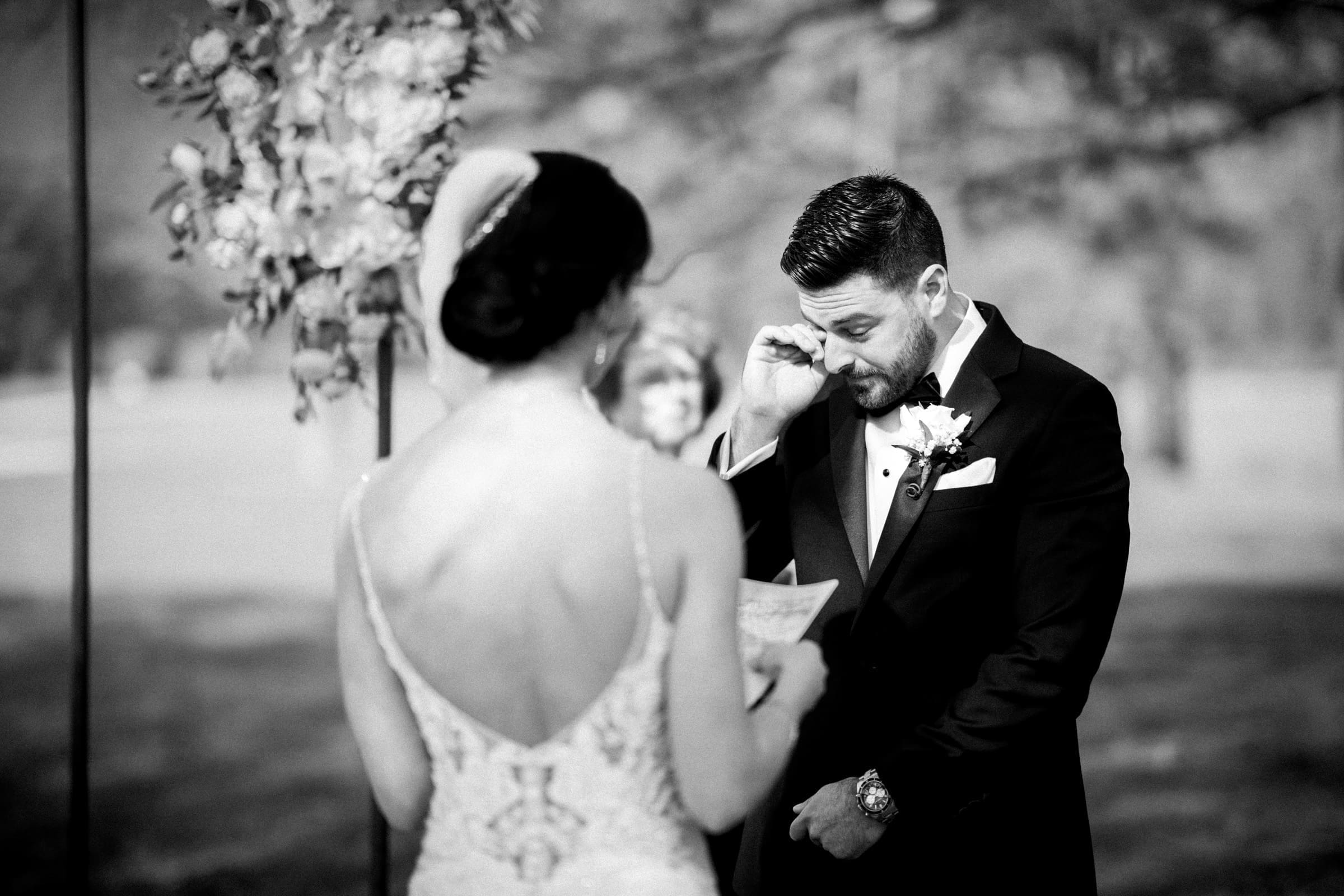 At the meadows elopement, a groom in a tuxedo wipes a tear during the intimate outdoor ceremony. The bride, in an elegant gown, stands opposite him holding a note. Flowers and blurred trees enhance the emotional scene.