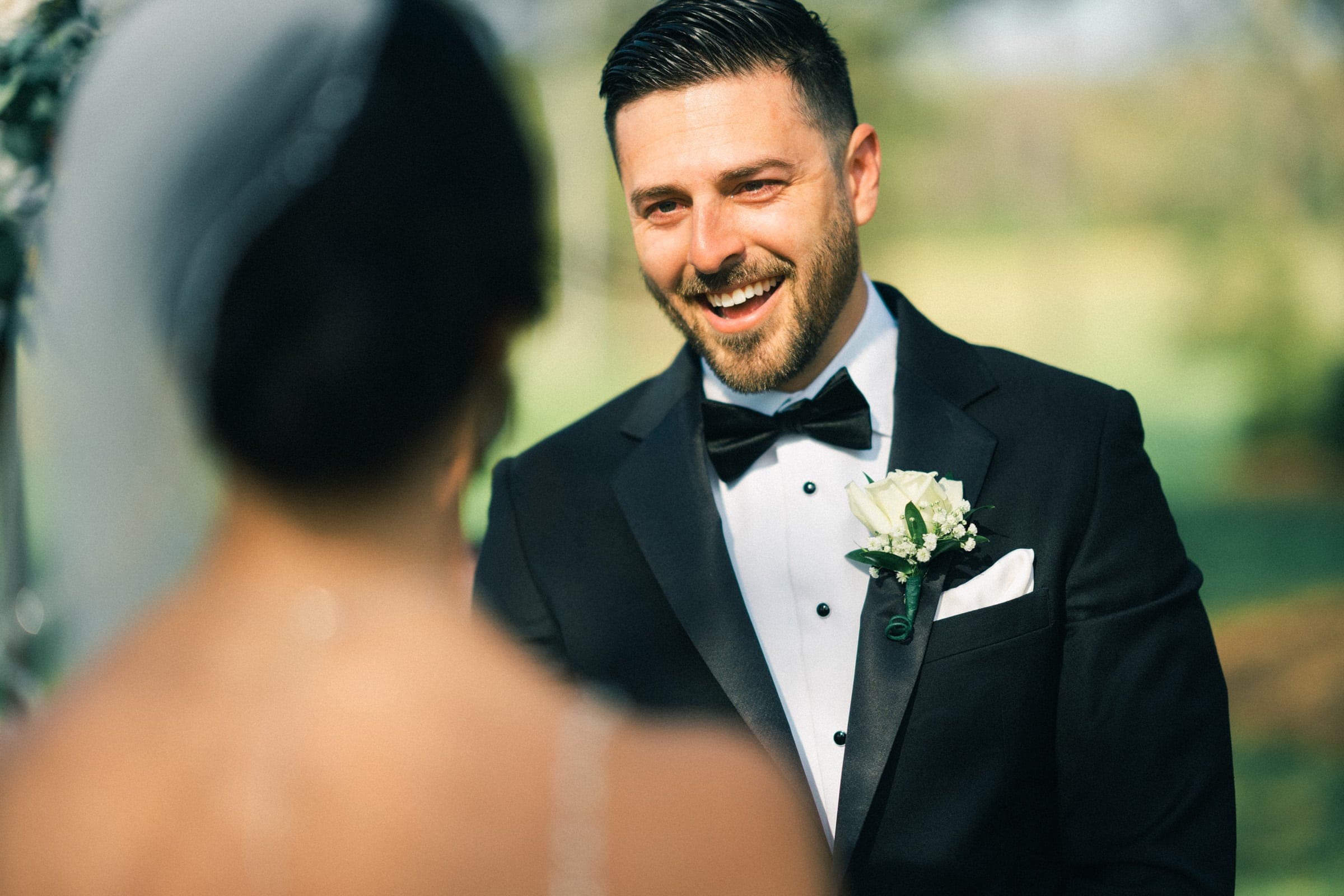 A groom in a black tuxedo smiles warmly at his bride during an elopement ceremony in the meadows. He wears a bow tie and a white rose boutonniere. The background is softly blurred, suggesting a sunny garden setting.
