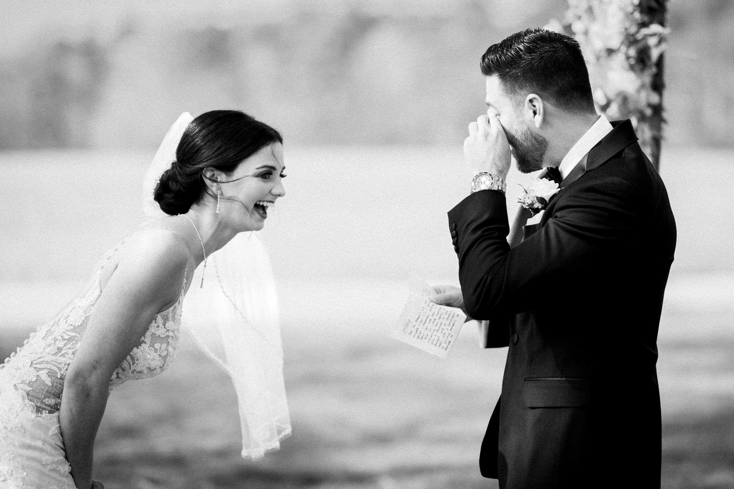 A bride and groom share a joyful moment during their meadows elopement. The bride leans forward, laughing, while the groom wipes away a tear, holding a piece of paper. This black-and-white scene captures an emotional and candid moment in the serene outdoor setting.