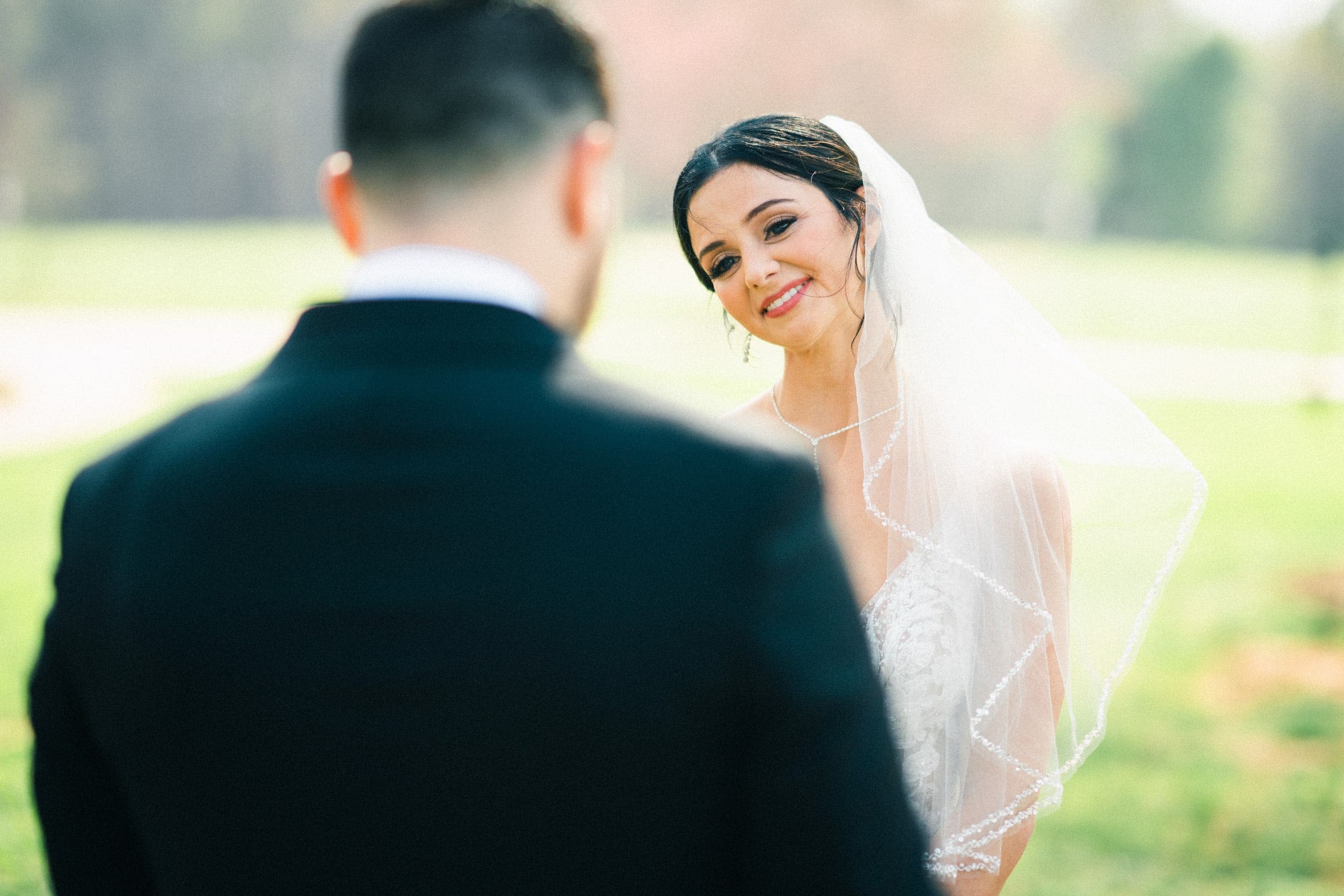 In a stunning lace dress and white veil, the bride smiles warmly at the groom silhouetted in his dark suit. The couple stands outdoors, embracing their intimate elopement amid the soft greenery of the meadows, bathed in natural sunlight.