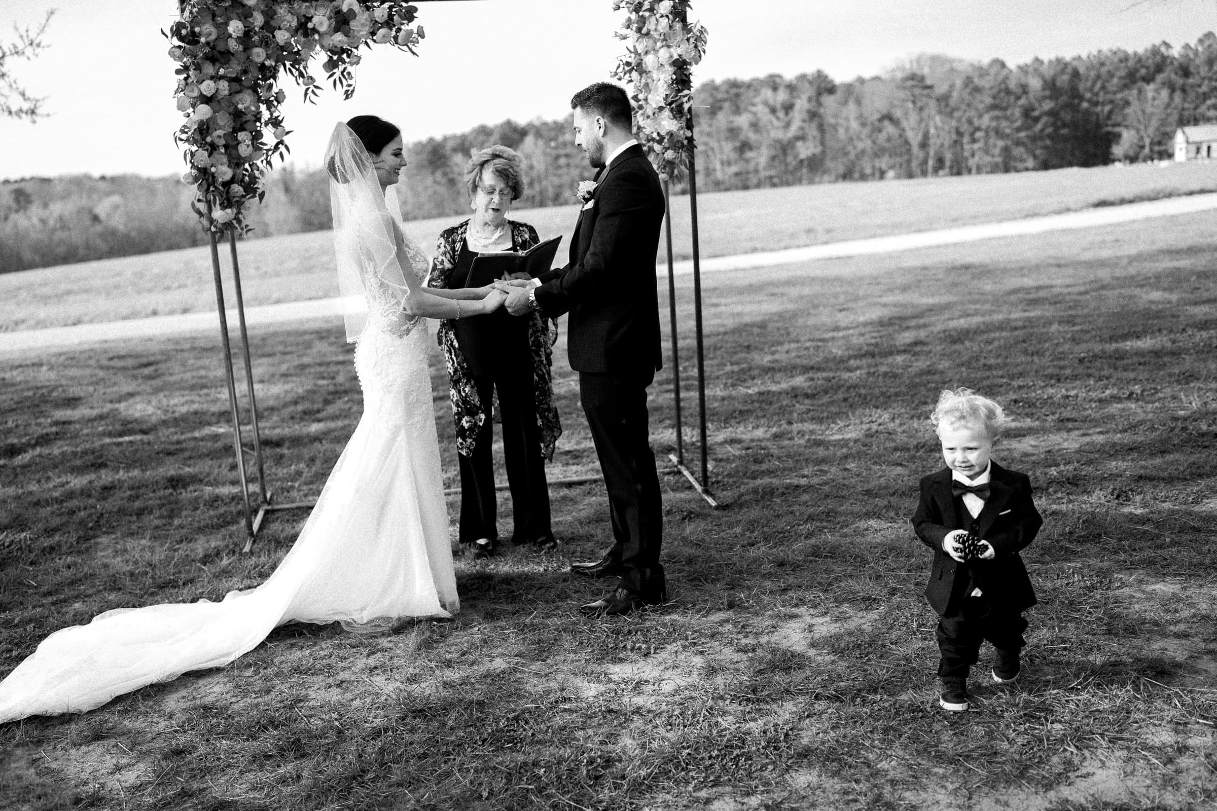 A bride and groom stand under a floral arch during their outdoor wedding ceremony at The Meadows Elopement, holding hands with an officiant present. In the foreground, a young child dressed in a suit playfully holds a small object, smiling broadly.
