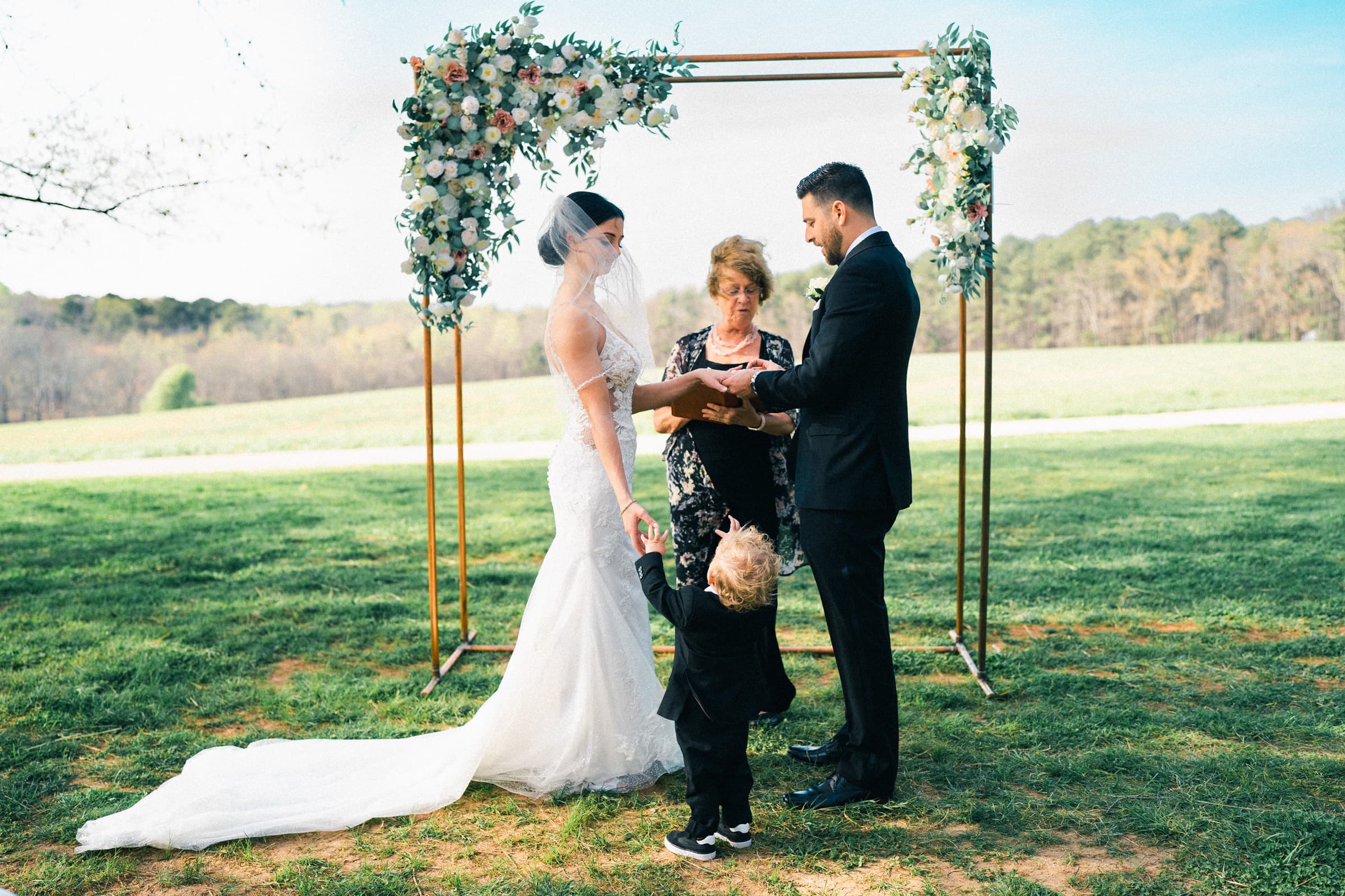 A bride and groom stand under a flower-adorned arch at their intimate elopement in the meadows. A young child gently holds the bride's hand, while a woman, possibly an officiant, reads from a book. The serene background features a grassy field and lush trees.