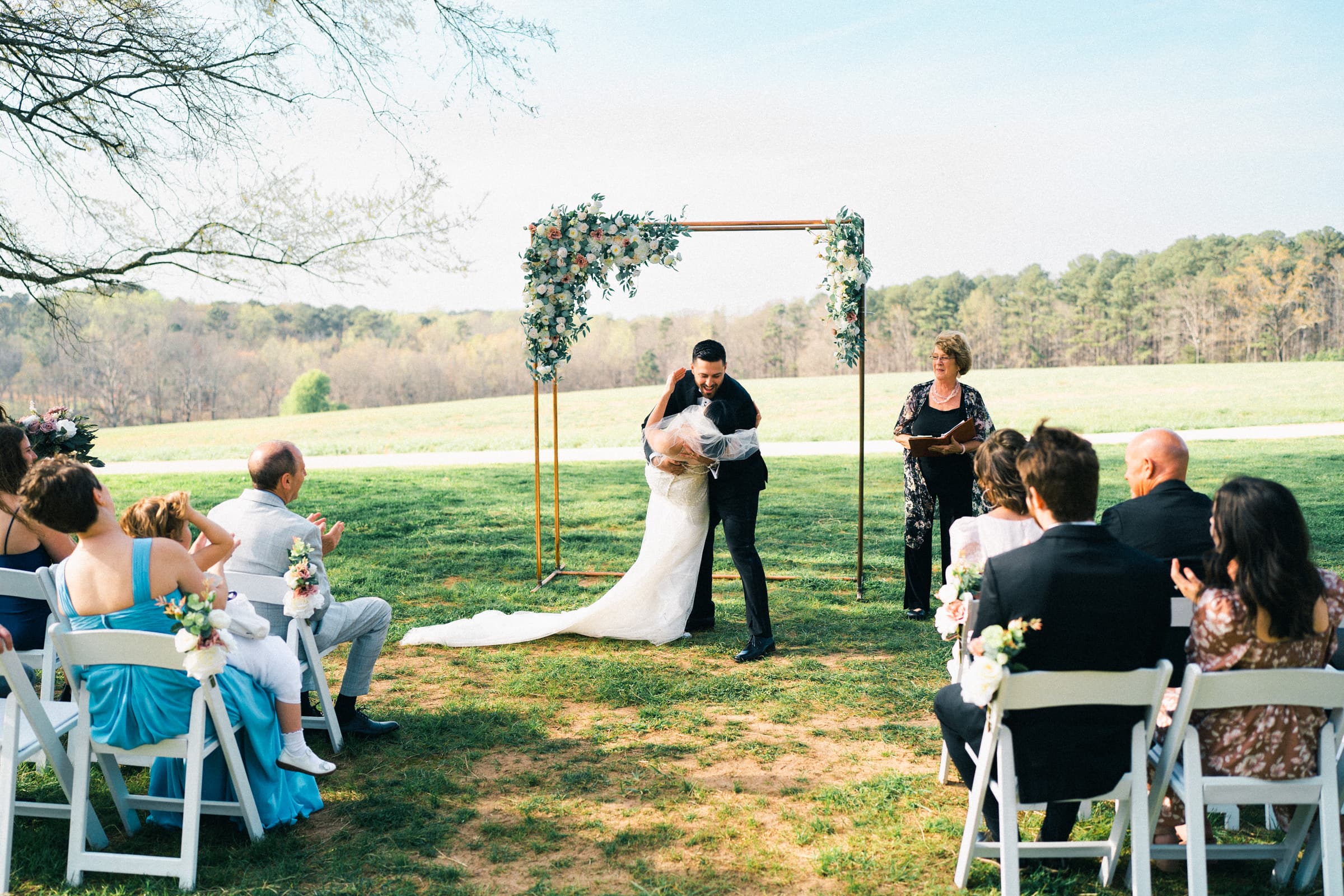 A bride and groom embrace under a floral arch during an outdoor wedding ceremony at The Meadows. Guests sit on white chairs, watching the couple. A woman stands beside them, likely the officiant. The intimate elopement unfolds on a grassy field with trees in the background.