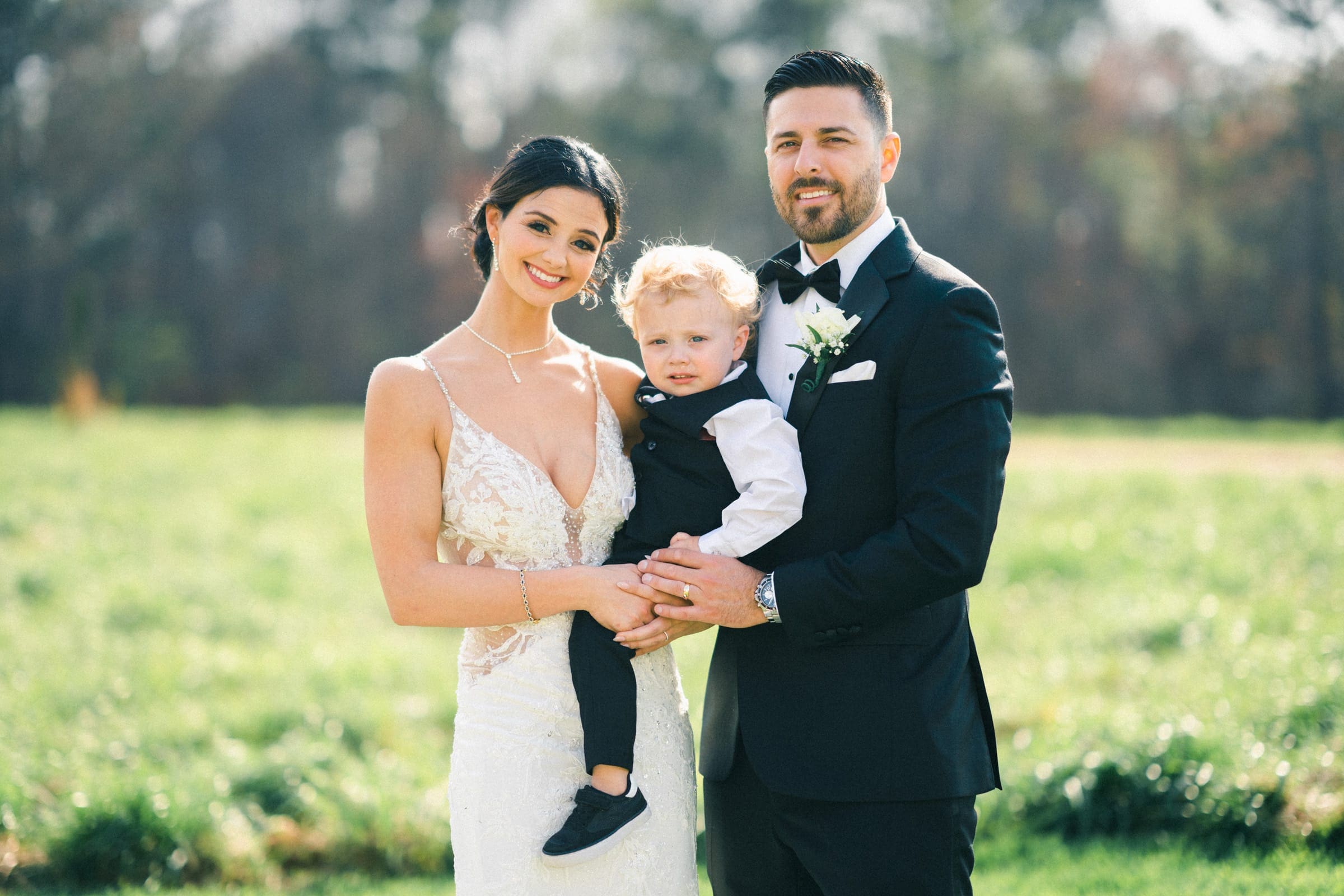 A couple in formal attire poses outdoors on a sunny day, holding a young child in their arms. The woman, wearing a white dress, and the man, dressed in a black suit, stand in the meadows. A grassy field and blurred trees are in the background.