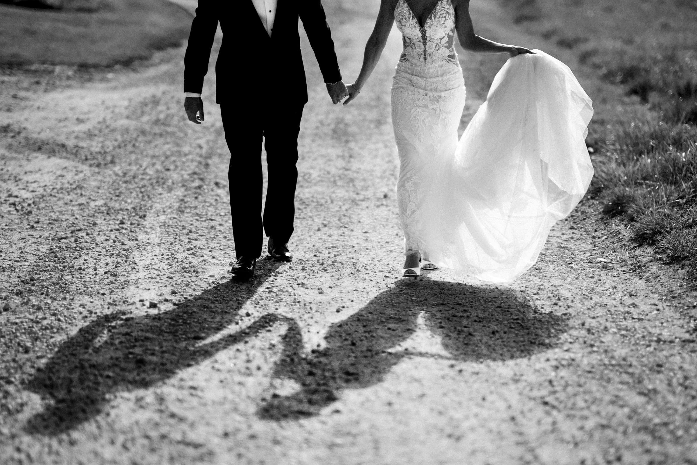 A newlywed couple in formal attire holds hands while walking down a rustic, sunlit path through the meadows. Their shadows stretch ahead, and the bride gracefully lifts her flowing white dress, capturing the essence of an intimate elopement.