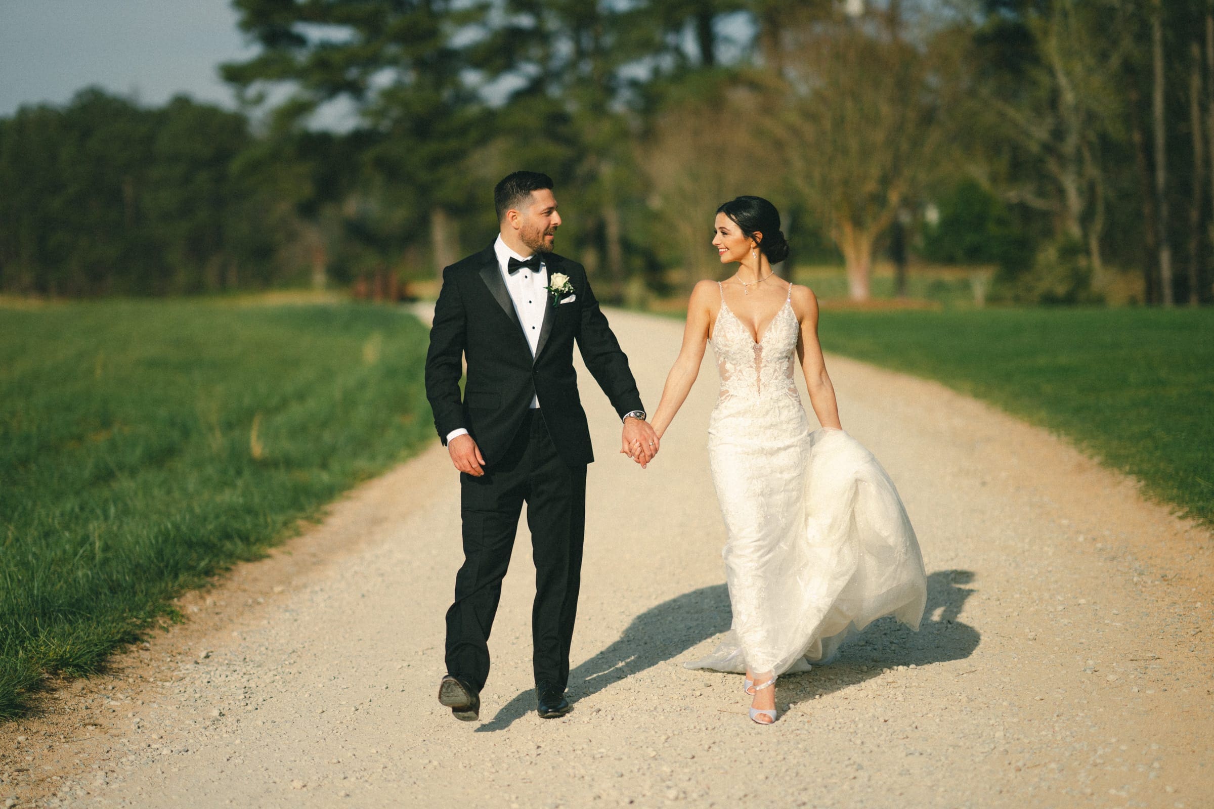 A bride and groom walk hand in hand along a tree-lined dirt path, celebrating their elopement. The bride wears a white dress, and the groom is in a black suit. Both are smiling amidst the greenery of the meadows under a clear sky.