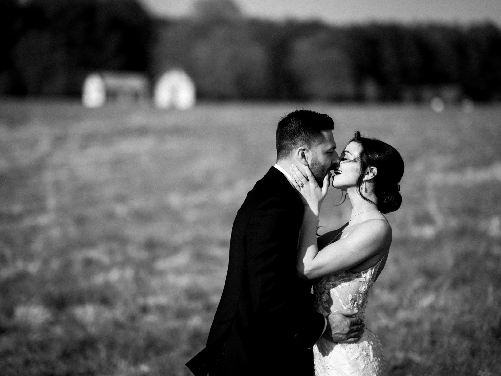 A black-and-white image captures a couple kissing in the meadows during their elopement. The bride wears a lace wedding dress, and the groom is in a dark suit. A blurred background with trees and a small building enhances the serene atmosphere.