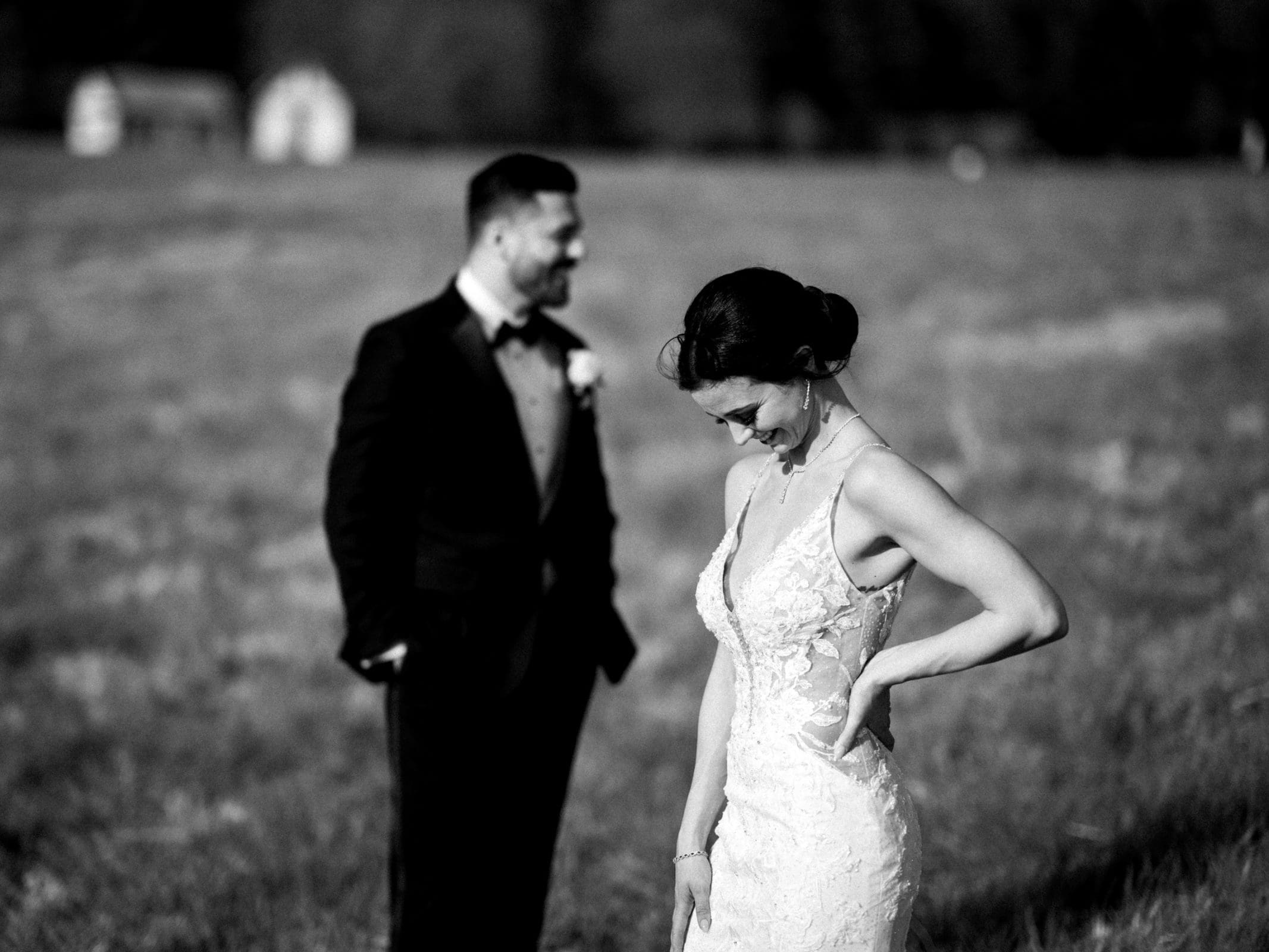 A bride in a detailed lace gown looks down, smiling shyly in the meadows. In the blurred background, a groom in a suit and bow tie watches her with a smile, capturing their elopement. The serene scene unfolds beautifully in black and white.