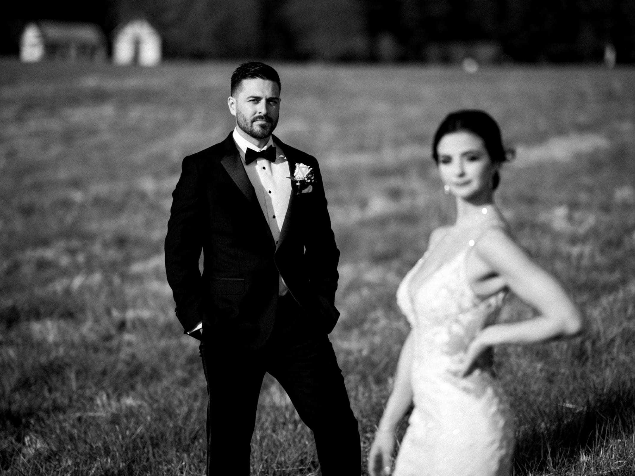 A black and white photo captures a groom in a tuxedo standing thoughtfully in the meadows, looking away. The blurred bride, ethereal in her wedding dress, stands prominently in the foreground. In the background, two small outbuildings add charm to this intimate elopement scene.