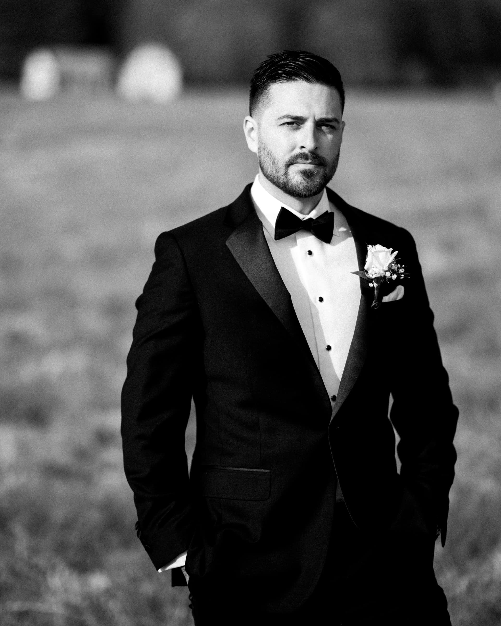 A man in a black tuxedo with a bow tie and boutonniere stands confidently in the meadows, hands in pockets, looking to the side. The blurred background enhances focus on him in this captivating black-and-white elopement photograph.