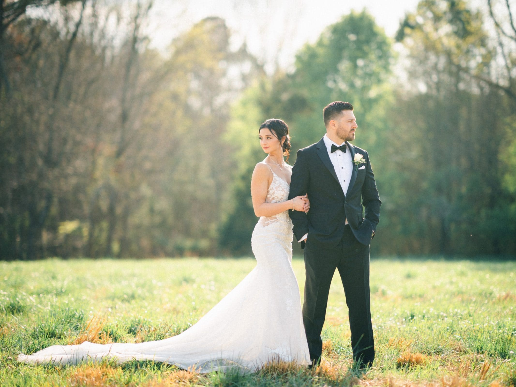 A bride and groom stand back to back in the sunlit meadows. The bride wears a long, white gown and the groom is in a black tuxedo. They are surrounded by lush greenery and trees, with soft sunlight filtering through the leaves—a perfect scene for an intimate elopement.