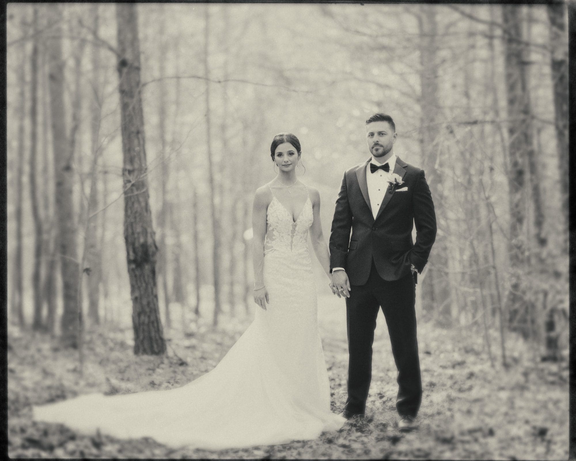 A black-and-white photo captures a couple during their meadows elopement. They stand hand in hand amidst the forest, framed by tall trees and a soft, blurred background. The bride's long white gown and the groom's suit with a bow tie add elegance to this intimate moment.