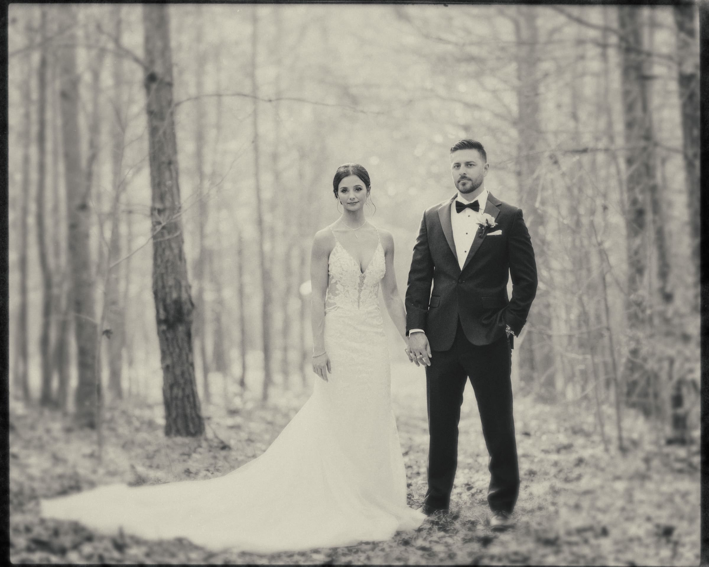 A black-and-white photo captures a couple during their meadows elopement. They stand hand in hand amidst the forest, framed by tall trees and a soft, blurred background. The bride's long white gown and the groom's suit with a bow tie add elegance to this intimate moment.