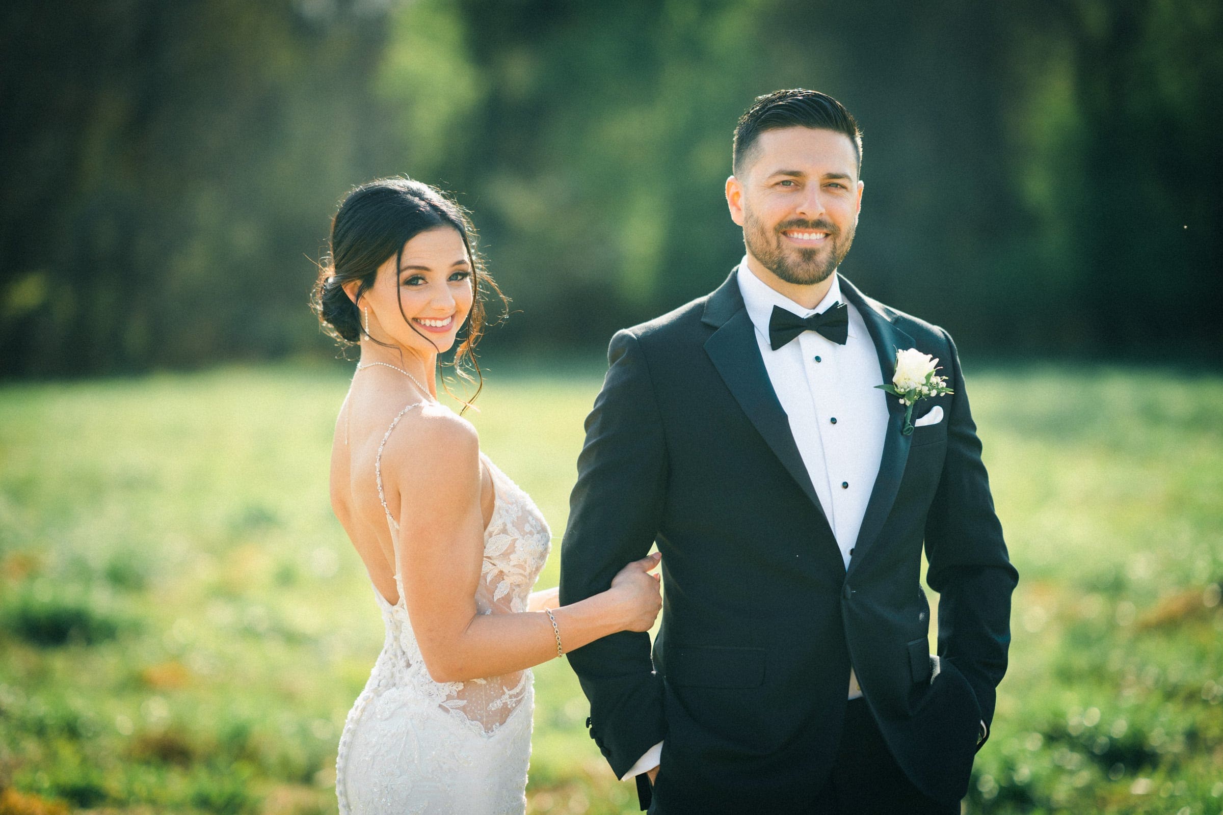 In the meadows, a couple stands together in formal attire. The woman in a white lace dress smiles and gently holds the man’s arm. The man, wearing a black tuxedo with a white boutonniere, beams beside her in this intimate elopement moment amidst blurred greenery.