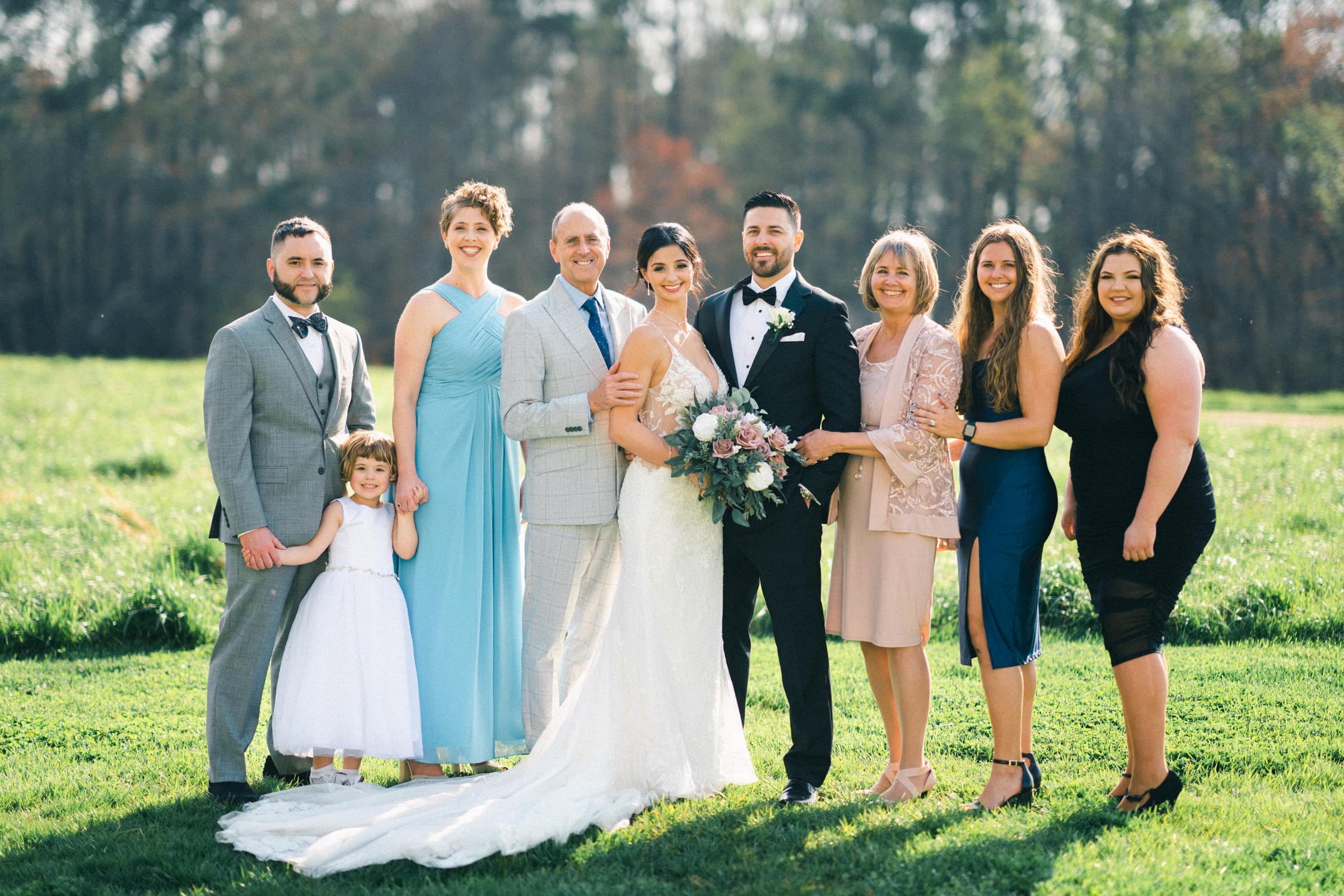 A newlywed couple stands in the center, surrounded by six adults and one child, all dressed in formal attire. They are posing for a photo outdoors on a sunny day with trees in the background at The Meadows. The bride holds a bouquet, capturing the spirit of an intimate elopement.