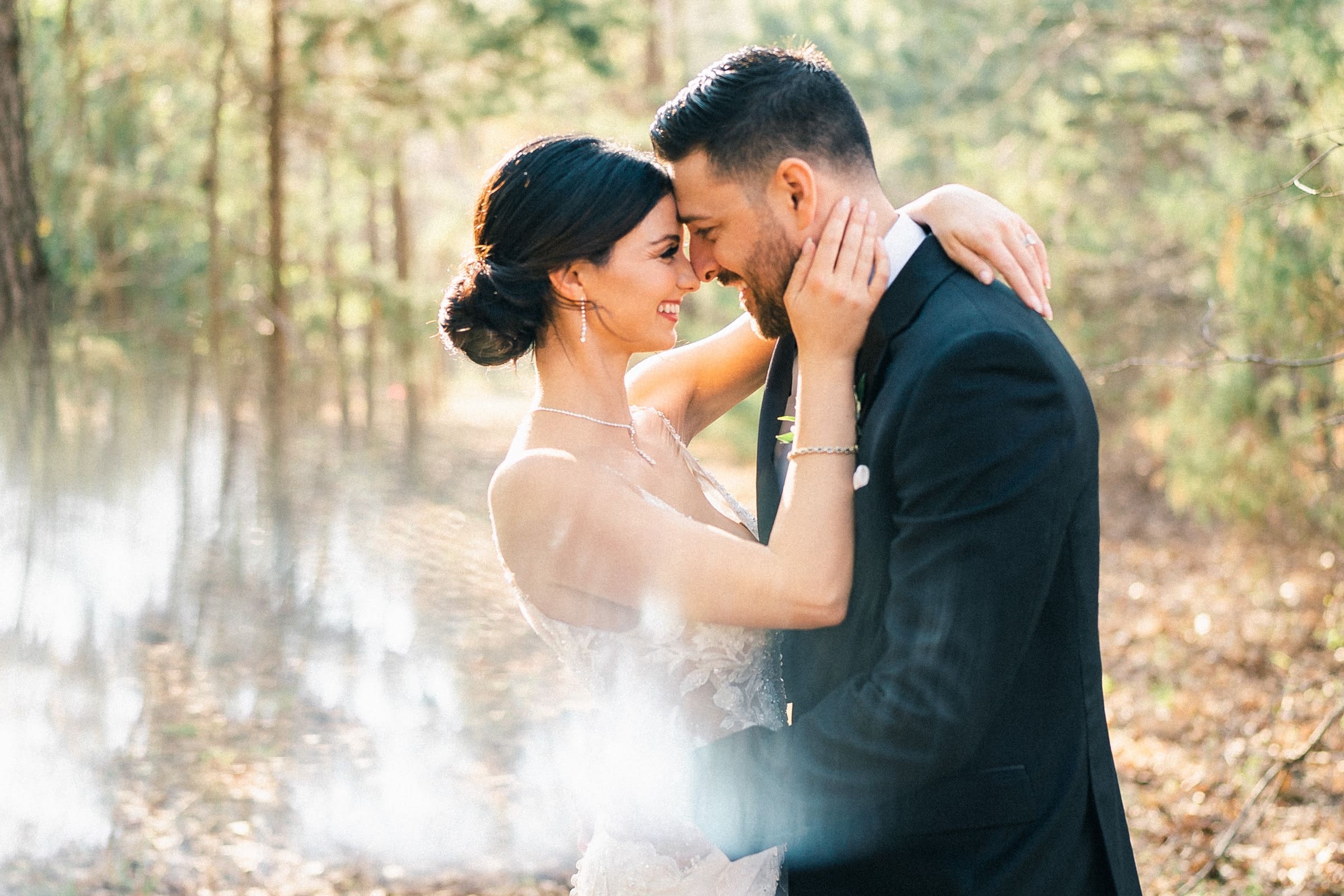 The bride and groom embrace, smiling at each other in a sunlit forest during their intimate elopement. She wears a white dress and he, a black suit. Sunlight filters through the trees of the meadows, crafting a softly romantic ambiance.