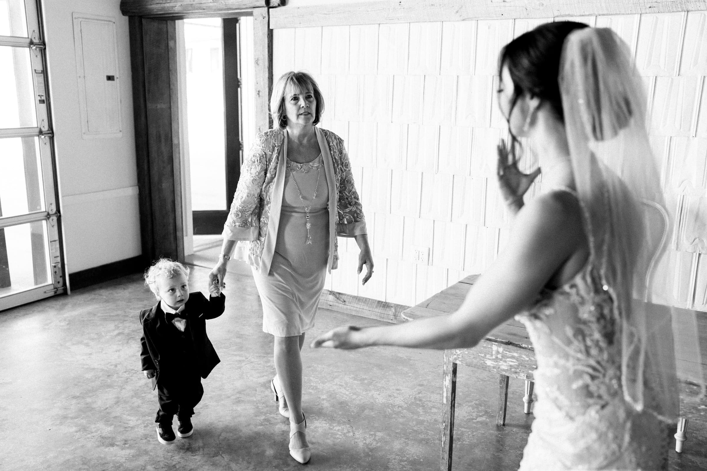 In the softly decorated room at The Meadows, a bride in a white dress and veil warmly greets an older woman holding a young boy's hand. The woman, dressed in a light-colored dress and lace jacket, smiles as the boy adjusts his suit jacket beside the rustic wooden table.