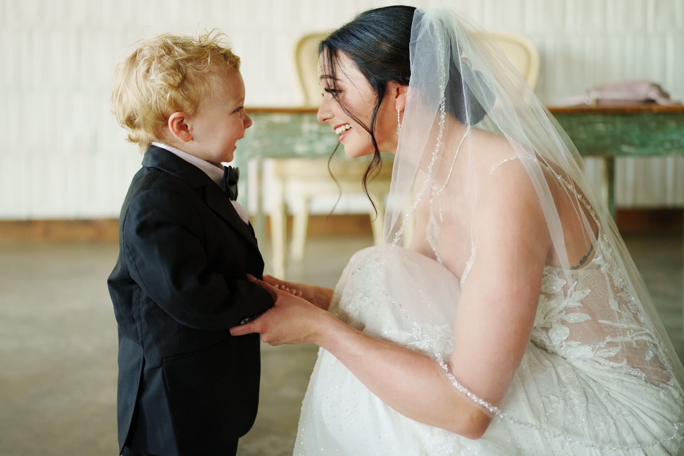 In a warmly lit room, a bride in a white wedding dress crouches down, smiling at a young boy in a black suit and bow tie. At their secret meadows elopement, they hold hands, sharing an unforgettable joyful moment together.