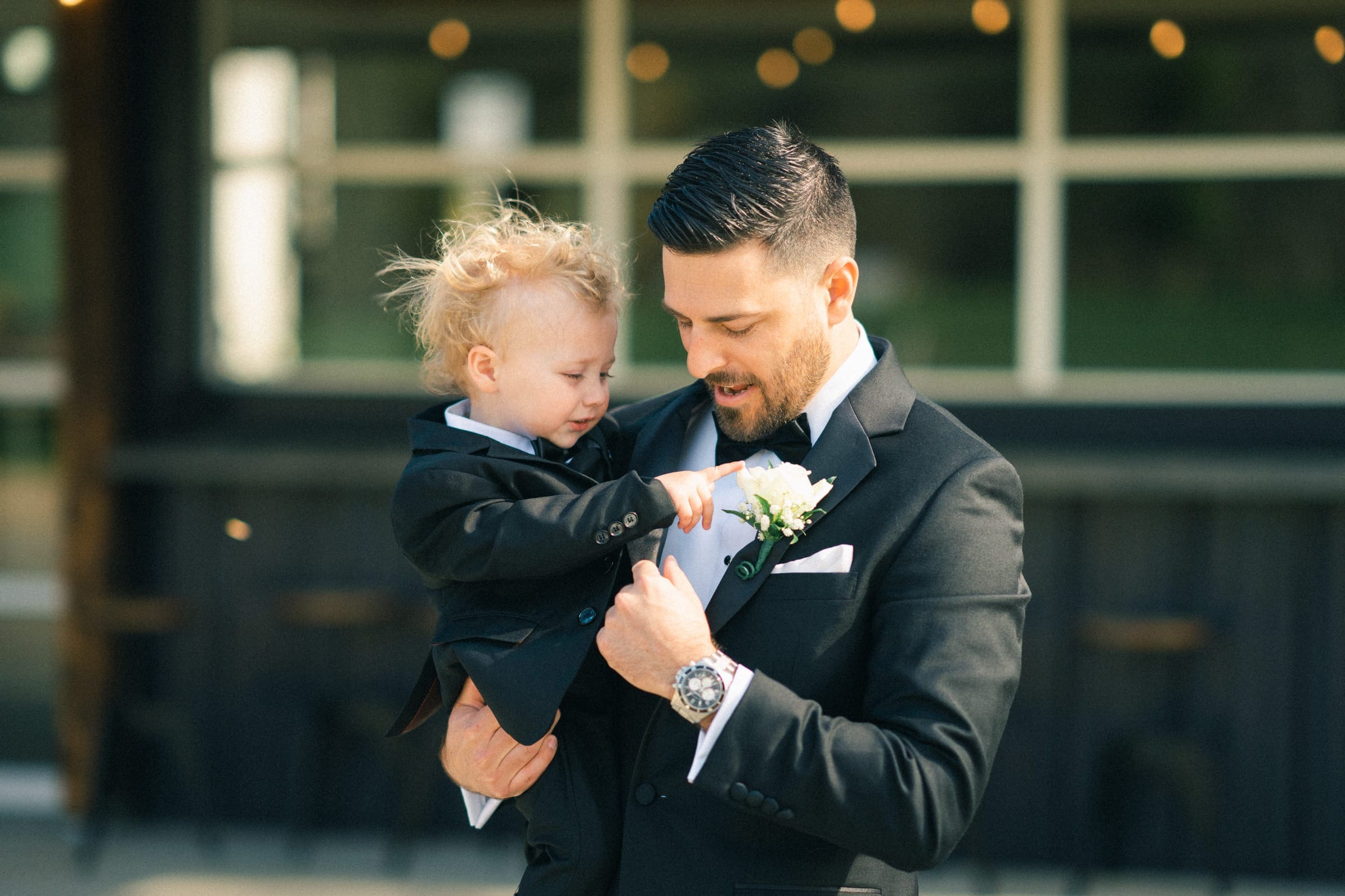 A man in a tuxedo holds a young child, both donning sharp attire, as they stand outdoors at The Meadows. The child curiously touches a white boutonniere on the man's jacket, their attention fixed. A blurred building forms the backdrop to this tender moment during their elopement.