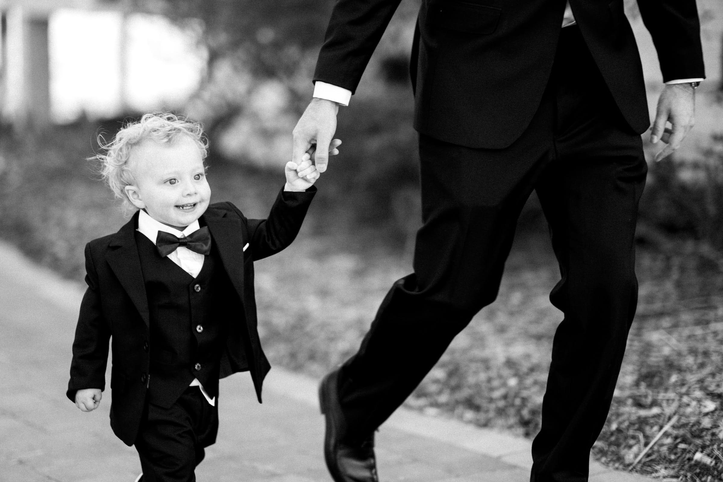 A smiling young child in a suit and bow tie holds the hand of an adult in a suit, strolling through the meadows. The scene is in black and white, capturing a joyful moment akin to an intimate elopement.