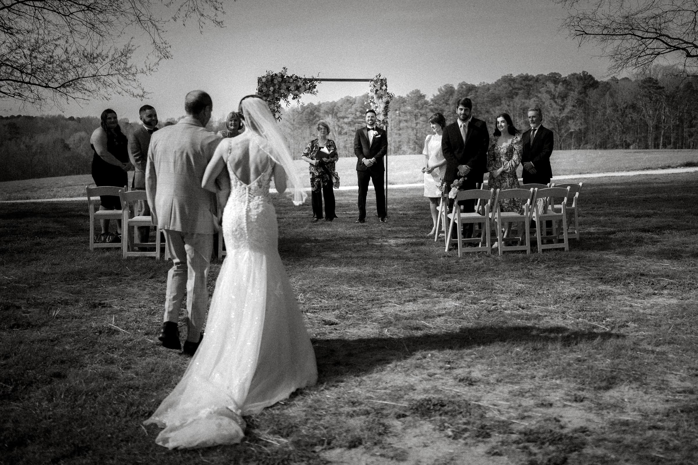 A black-and-white image captures an outdoor wedding ceremony in the meadows. A bride in a long gown walks down the aisle with a man during an intimate elopement. Guests are seated on either side, and the officiant awaits under a floral arch. Trees and fields grace the serene background.