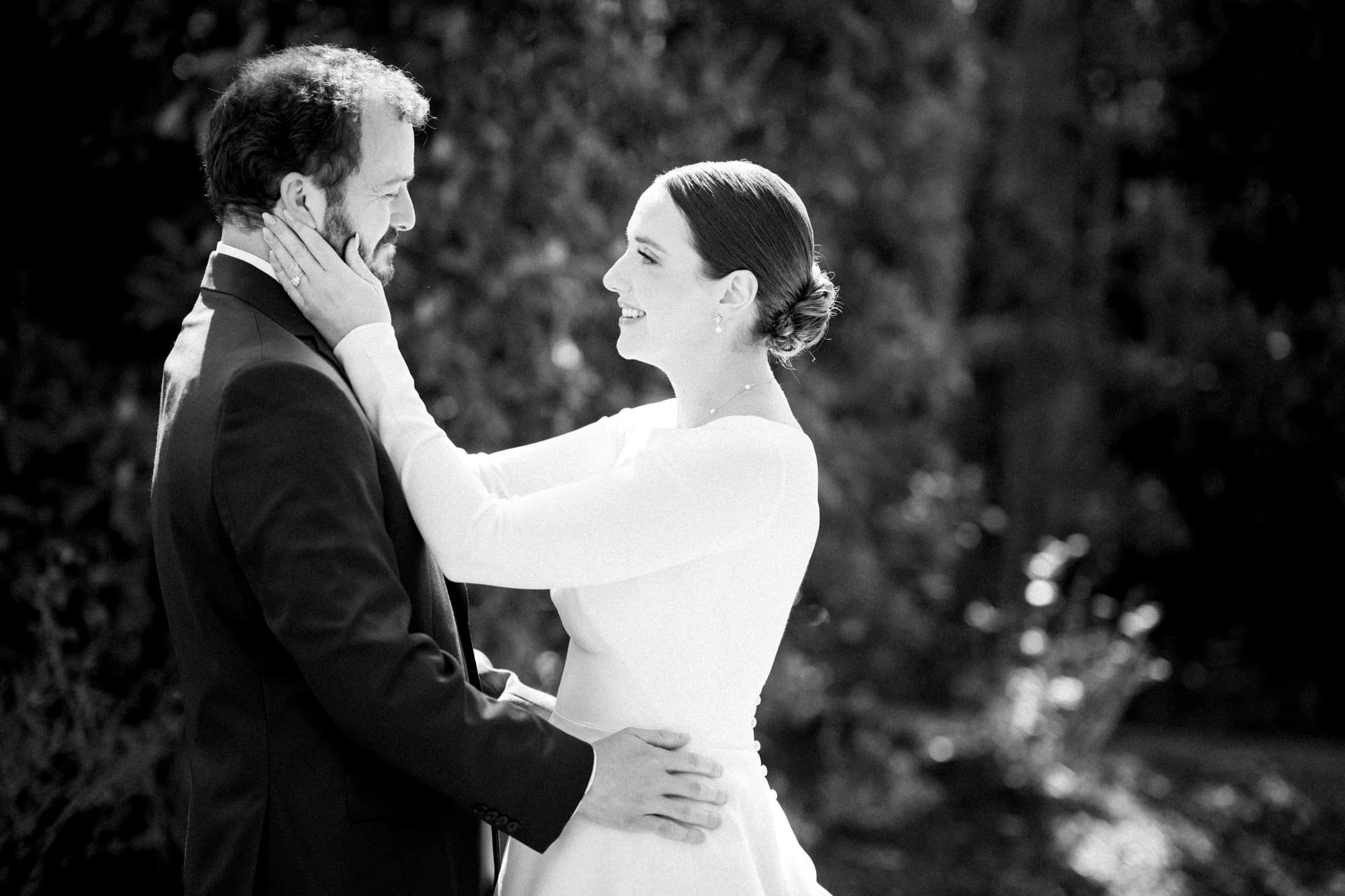 sutherland estate wedding - stephanie & hunter A black and white image captures a couple on their wedding day at Sutherland Estate. The bride, in a long-sleeved gown, holds the groom's face with both hands, smiling. He's in a suit, returning her gaze with a gentle smile. They're outdoors, framed by the estate's enchanting trees.