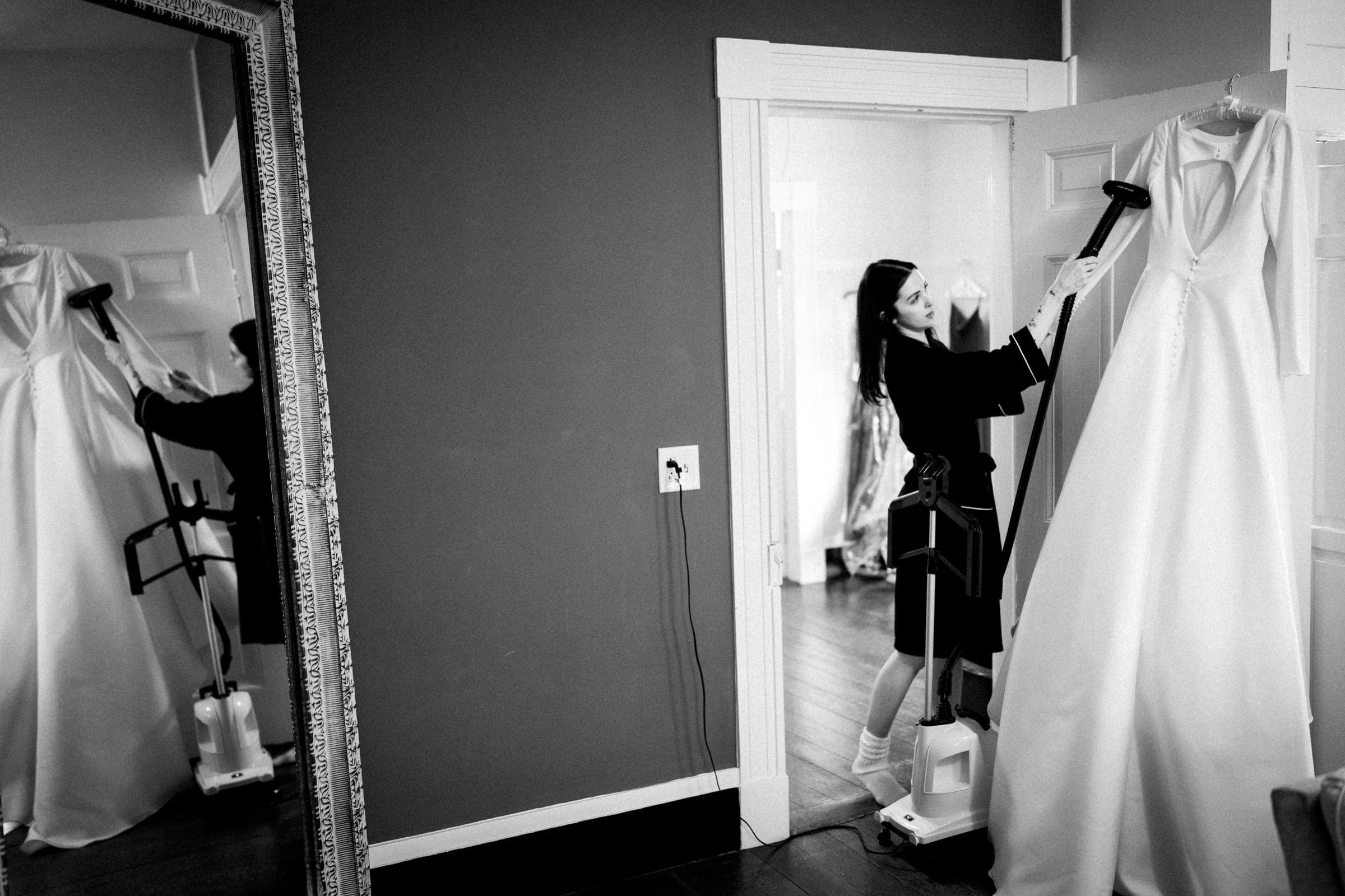 sutherland estate wedding - stephanie & hunter At a Sutherland Estate wedding, a woman steams a bridal gown hanging on a door in a room with a large mirror. The black-and-white image captures the dress's reflection and steam rising as she works, set against dark walls and wooden flooring.