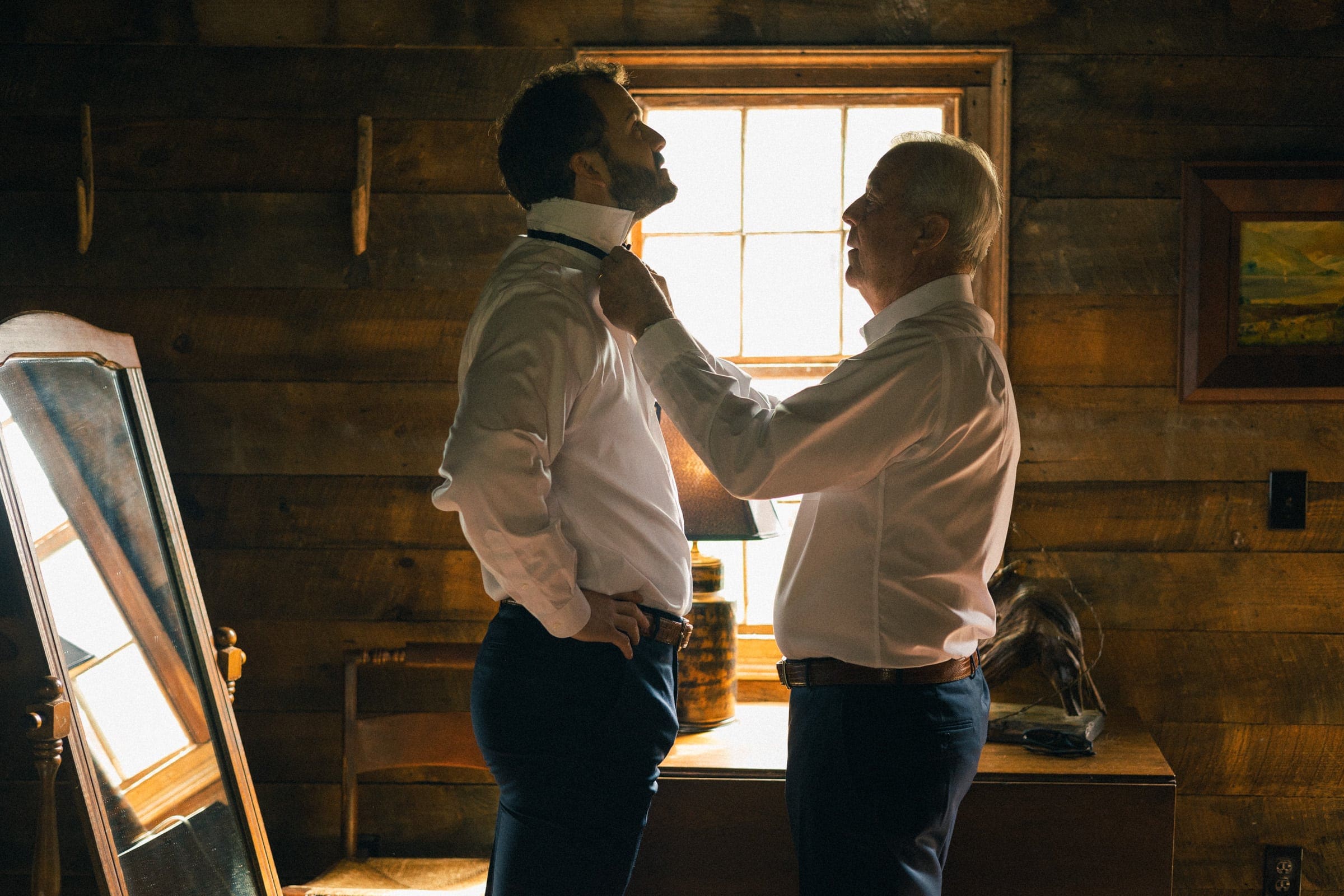 sutherland estate wedding - stephanie & hunter In a rustic room at Sutherland Estate, a man helps another with a bow tie. They stand near a window that casts warm light on them, illuminating the cozy, intimate atmosphere enhanced by a mirror and wooden furniture.