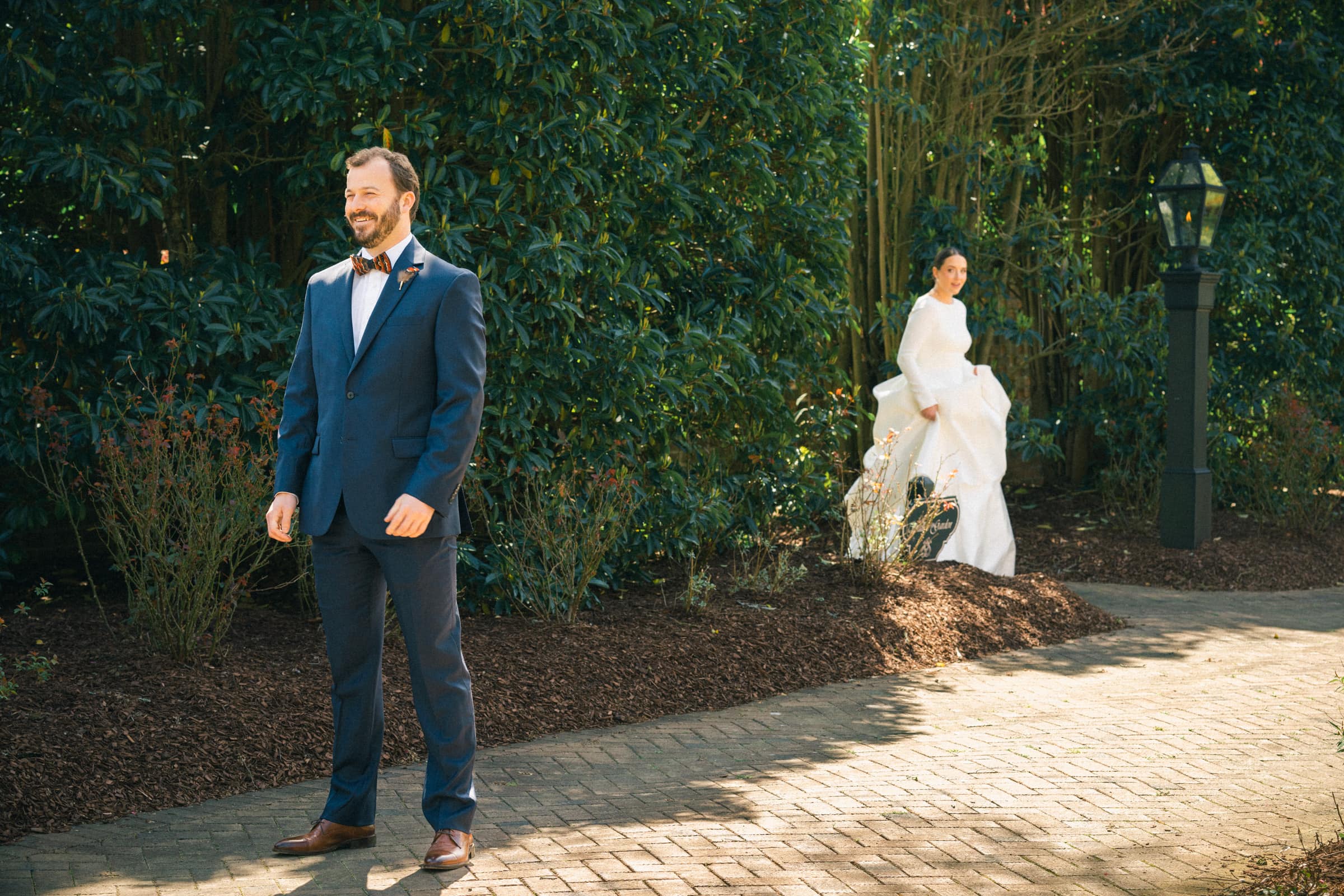 sutherland estate wedding - stephanie & hunter At a Sutherland Estate wedding, a groom in a blue suit stands smiling on a brick pathway. Behind him, hidden partially by bushes, the bride in her white dress approaches with her bouquet. A lamppost peeks through the lush greenery to the right.