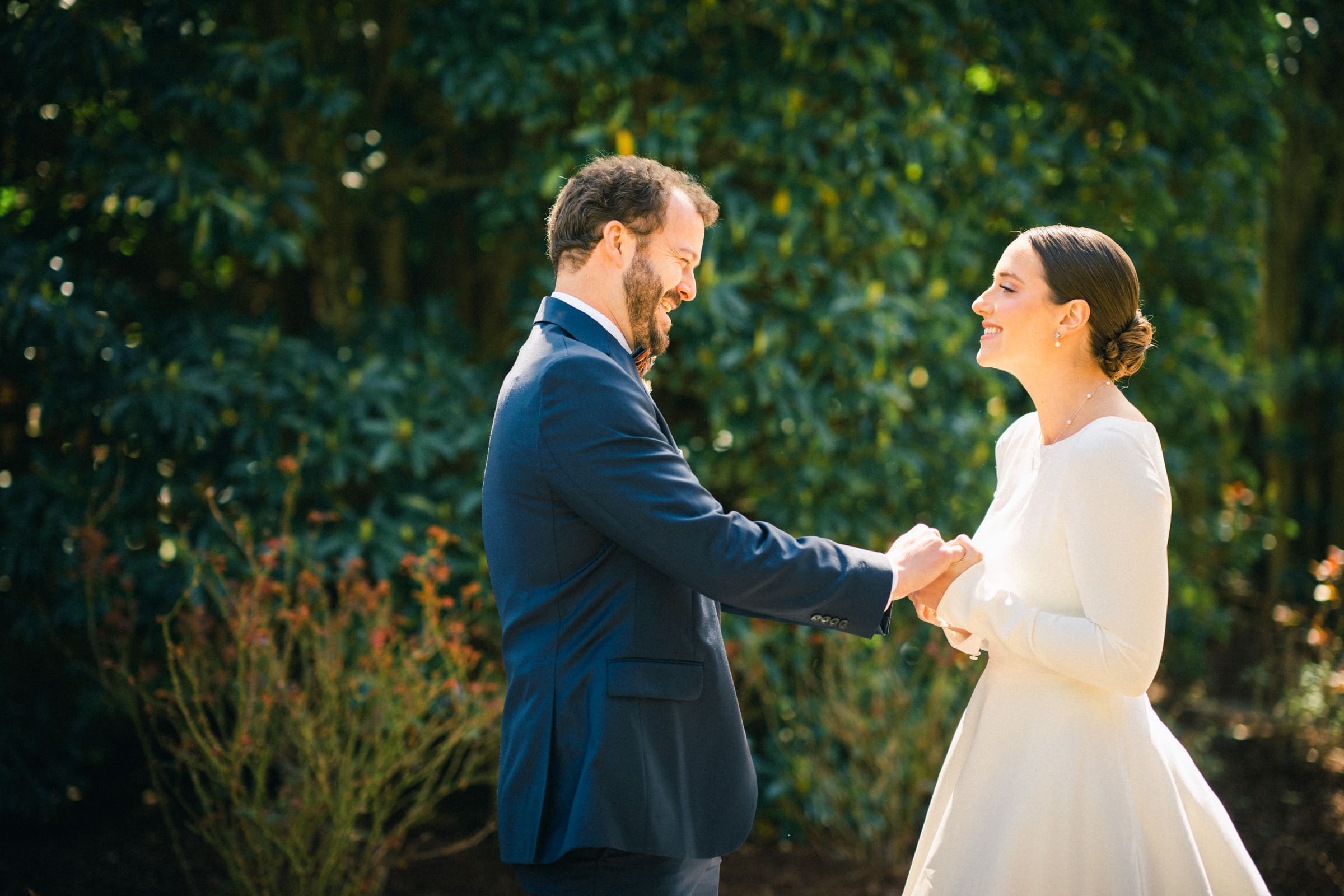 sutherland estate wedding - stephanie & hunter At a picturesque Sutherland Estate wedding, a couple dressed in formal attire stands facing each other outdoors. The man, in a navy suit, holds the hands of the woman in a white dress. Both are smiling, surrounded by lush greenery.