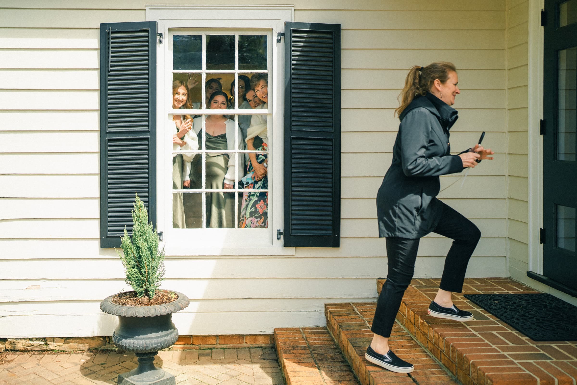sutherland estate wedding - stephanie & hunter A woman in a black jacket and pants steps up the brick stairs of Sutherland Estate, phone in hand. Inside, four women smile and wave from a window, framed by the light yellow house with black shutters. A potted plant adds charm to this perfect wedding venue entrance.