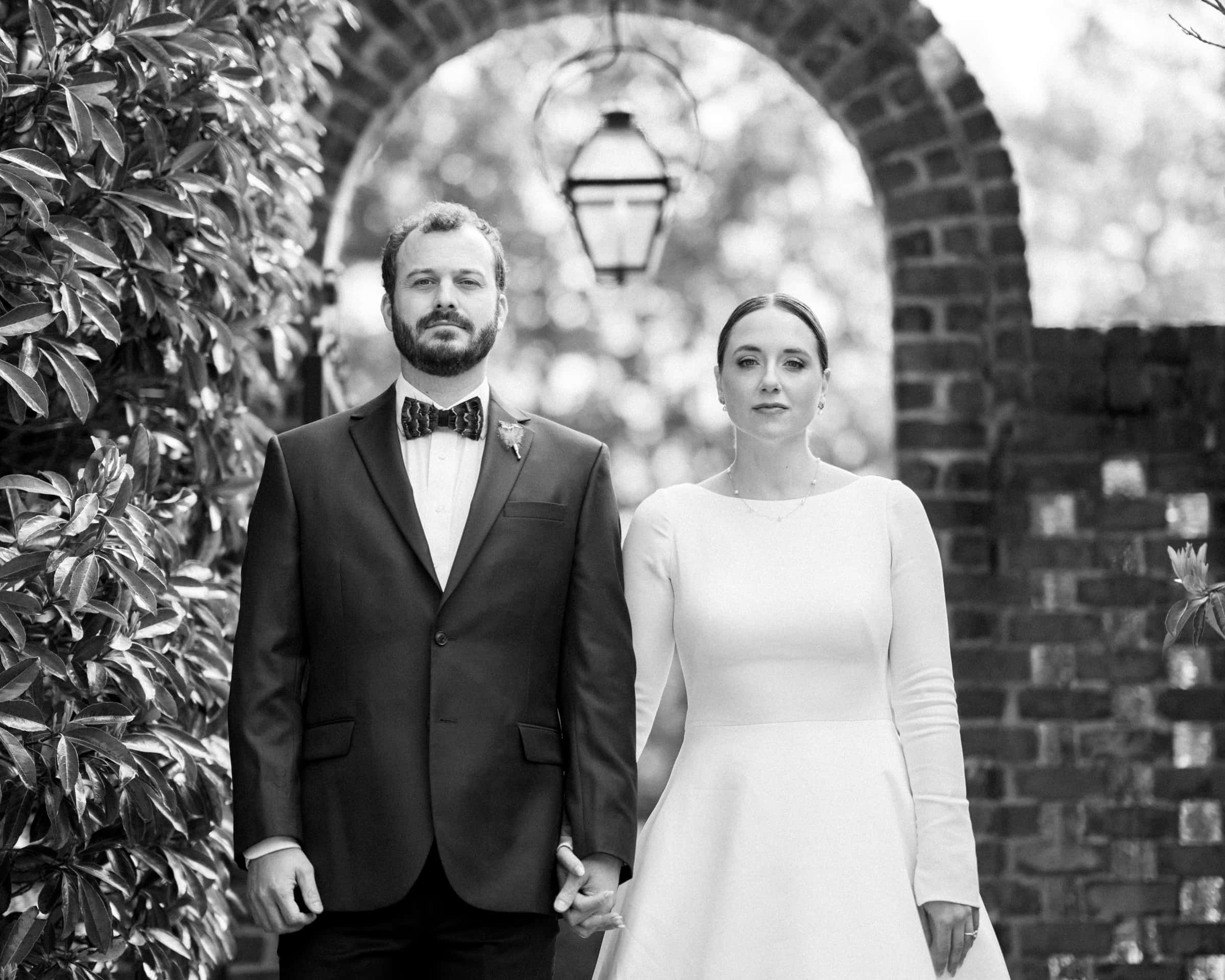 A timeless black and white photo captures a couple holding hands at their Sutherland Estate wedding. The man is in a suit with a bow tie, the woman in a white dress. They stand before a brick archway adorned with a lantern, flanked by lush foliage.