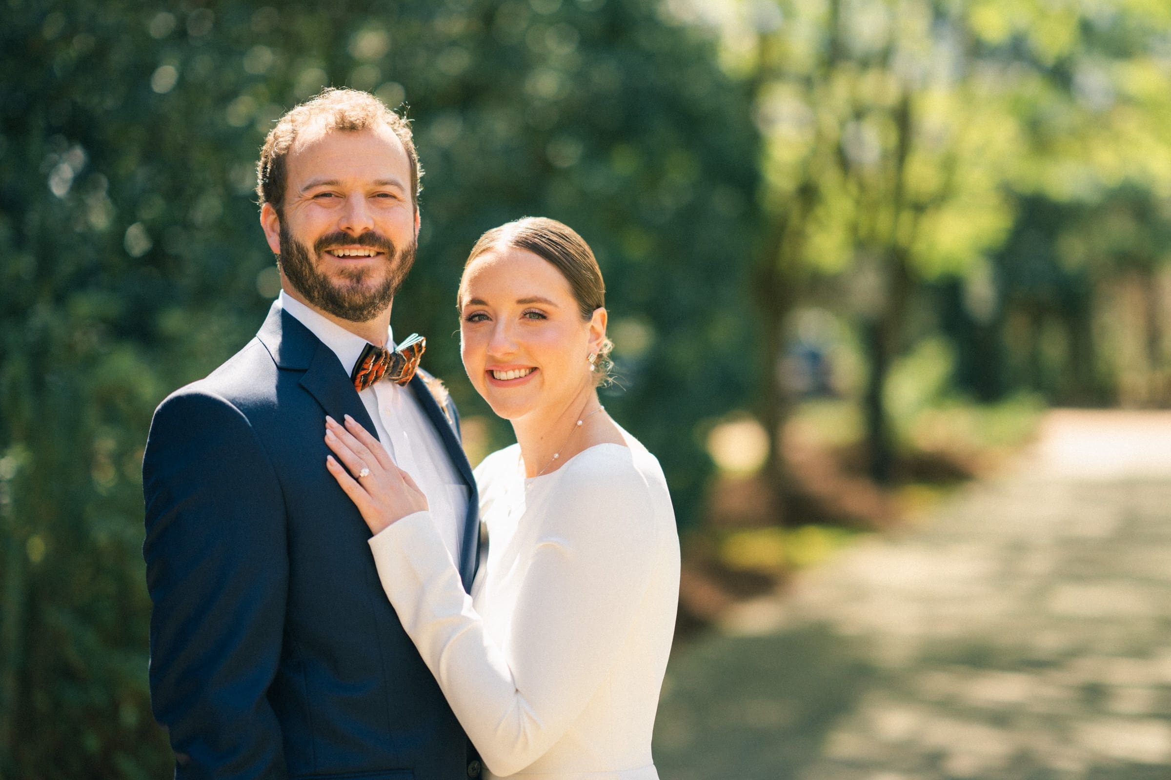 sutherland estate wedding - stephanie & hunter A couple in formal attire beams amid the lush greenery of Sutherland Estate on a sunny day. The woman dazzles in a white dress, while the man complements her perfectly in a navy suit with a bow tie.