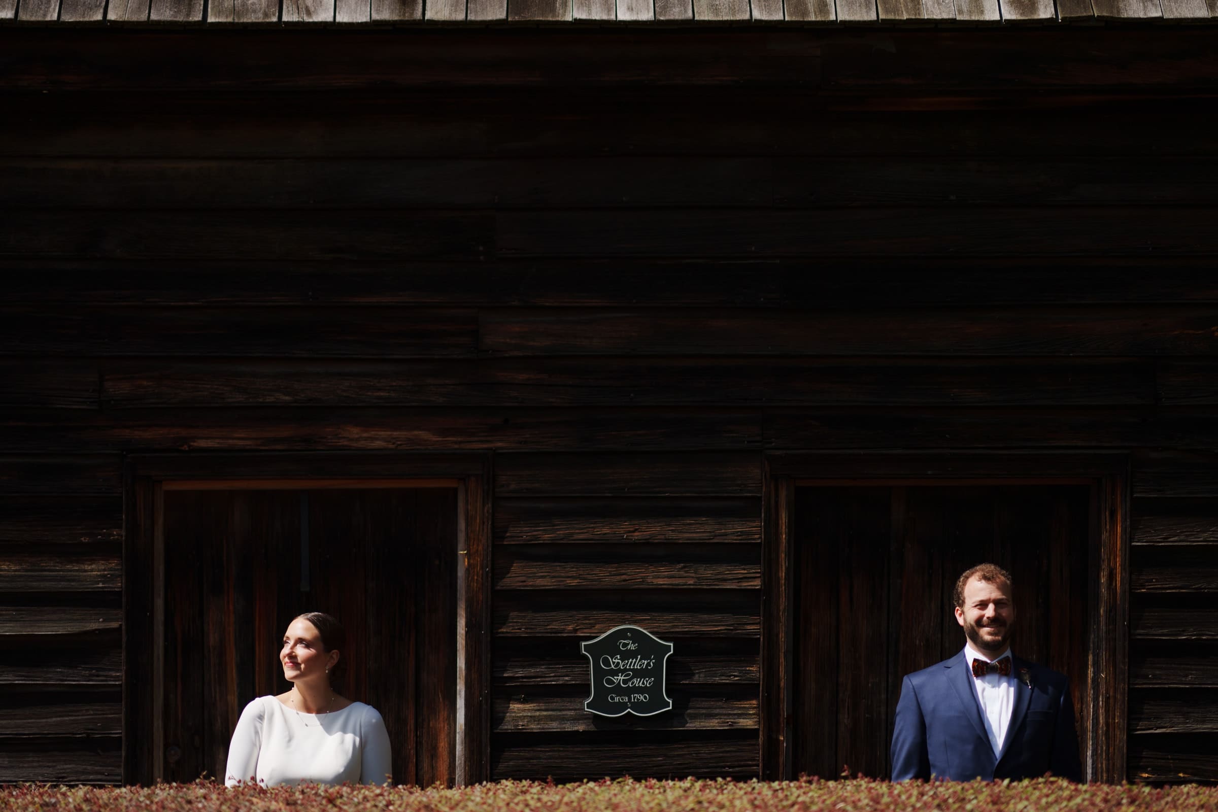 sutherland estate wedding - stephanie & hunter A woman and a man stand in separate doorways of the dark wooden building, exchanging smiles at Sutherland Estate. The plaque between them reads The October House, Circa 1780.