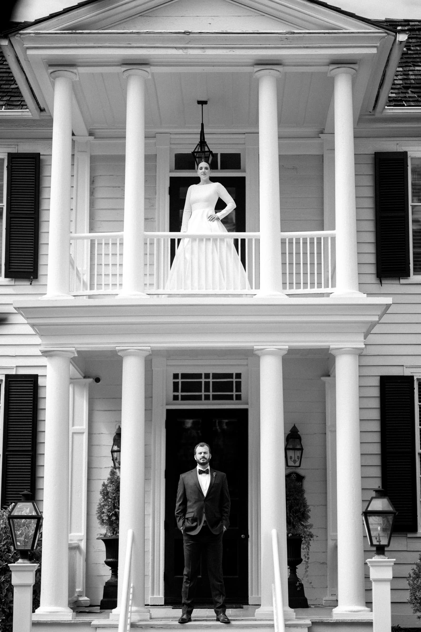 sutherland estate wedding - stephanie & hunter A bride stands on a second-floor balcony in a long white dress at the Sutherland Estate, while a groom in a suit poses on the front steps of the grand, white-paneled house with columns. The black-and-white image captures an elegant, timeless wedding moment.