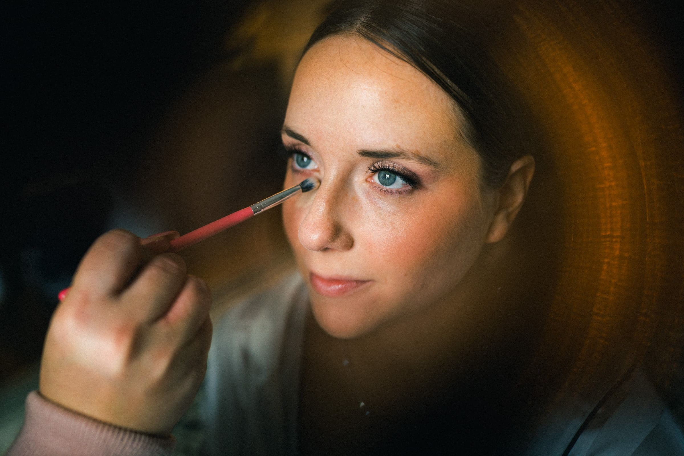 sutherland estate wedding - stephanie & hunter A person with blue eyes is having makeup applied with a brush to their upper eyelid, preparing for a magical Sutherland Estate wedding. The lighting creates a warm, circular glow around them, beautifully highlighting the focus on their face and the intricate makeup process.