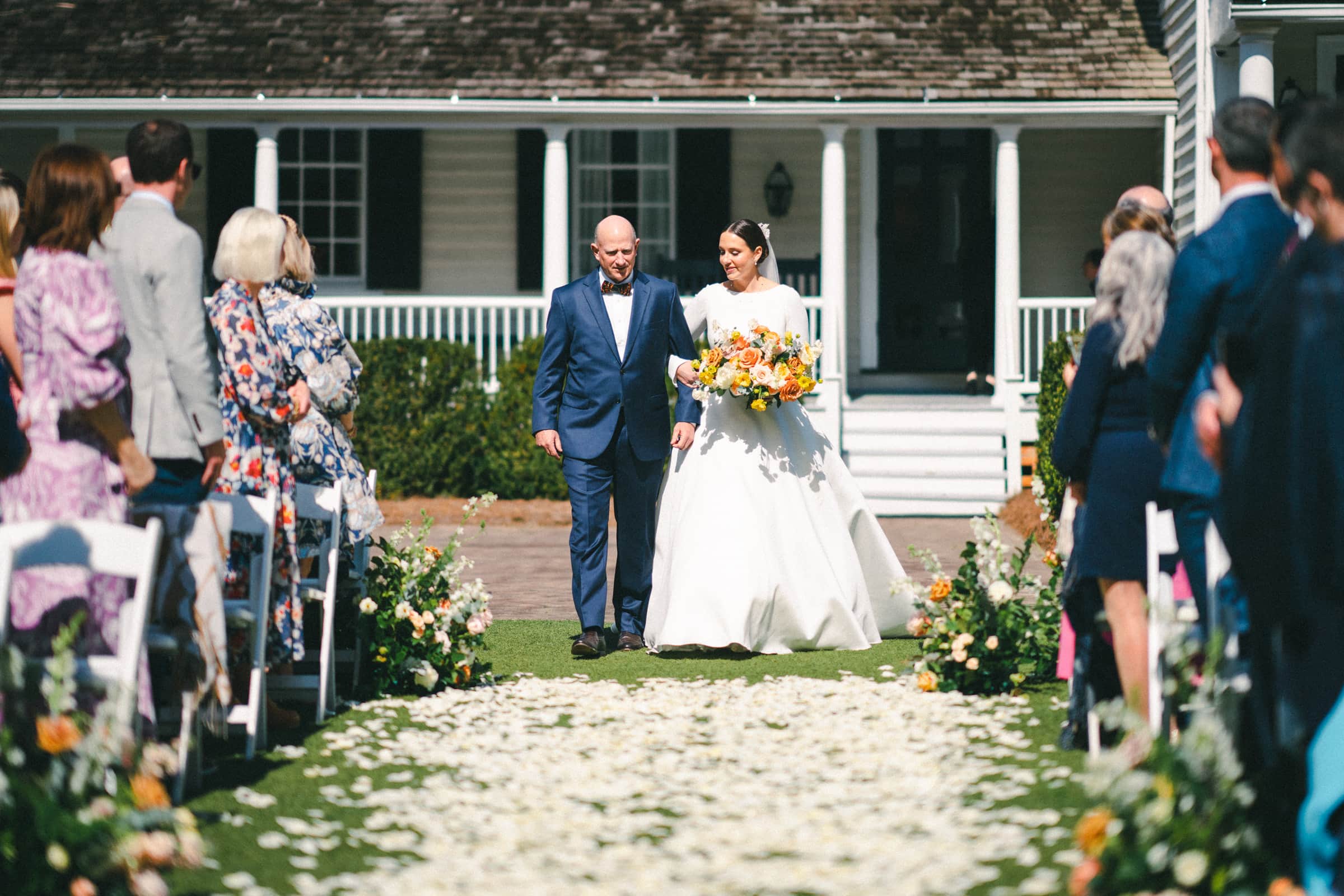 sutherland estate wedding - stephanie & hunter At the picturesque Sutherland Estate wedding, a bride and an elderly man, possibly her father, gracefully walk down the aisle during an outdoor ceremony. She holds a bouquet of flowers as guests on either side look on. In the background, a charming house with a porch completes this heartfelt scene.