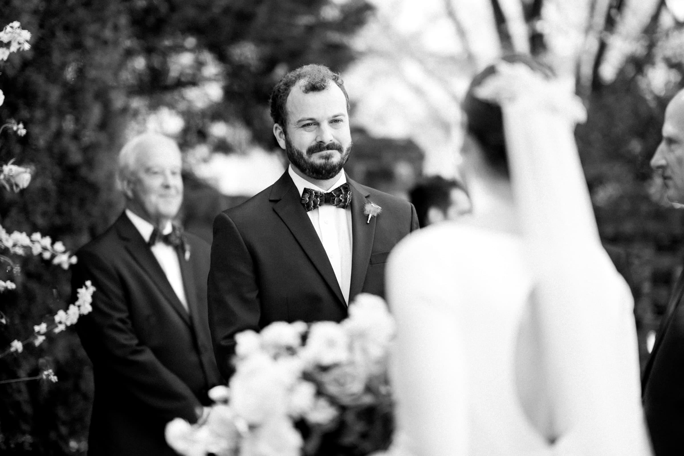 sutherland estate wedding - stephanie & hunter A black and white photo captures a groom in a suit with a bow tie at an elegant Sutherland Estate wedding. He faces the bride, blurred in the foreground, as two older men stand behind him. Flowers beautifully adorn the scene.