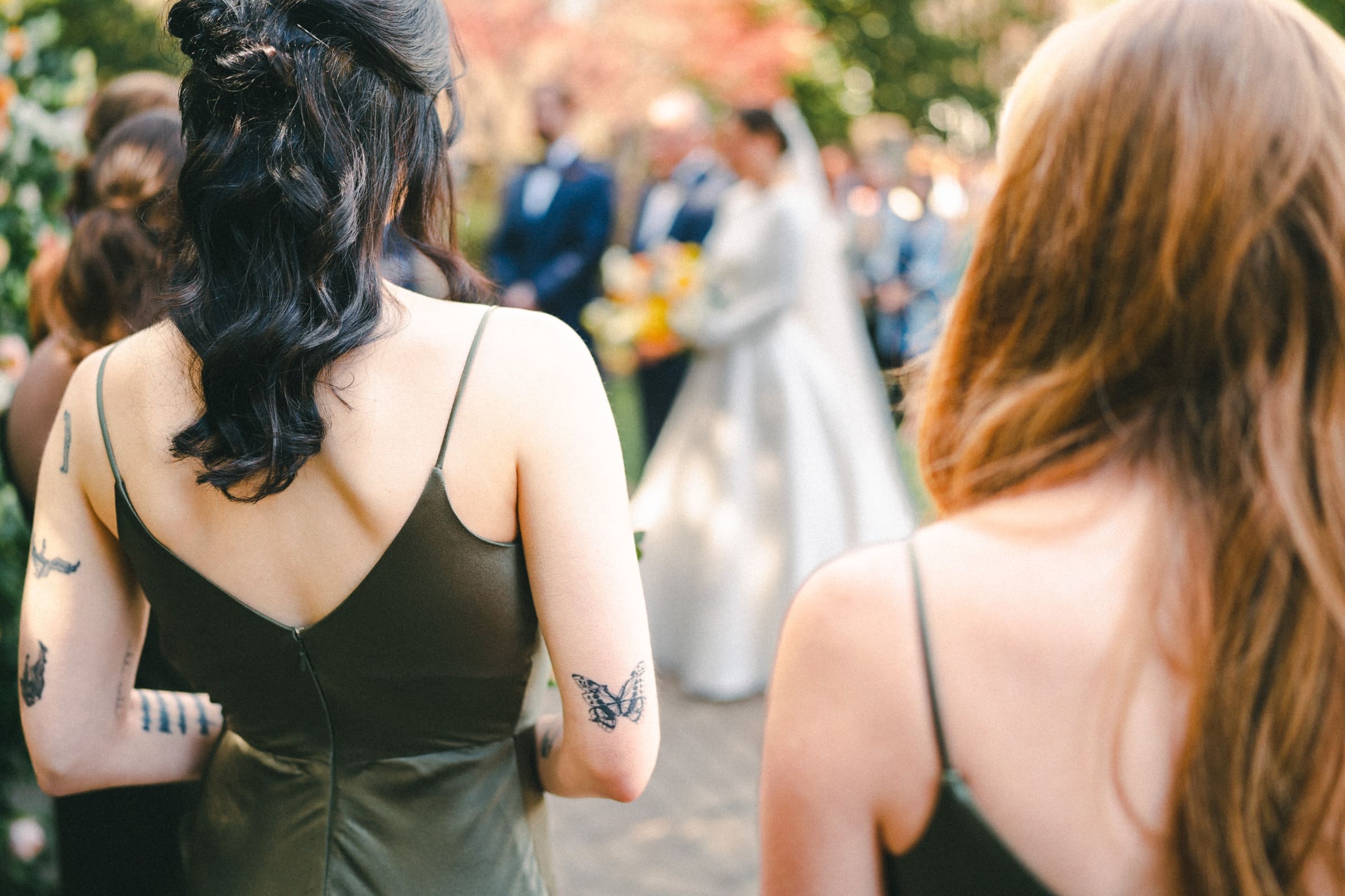 sutherland estate wedding - stephanie & hunter Two bridesmaids with butterfly tattoos on their arms stand in focus, wearing olive dresses, at a picturesque Sutherland Estate wedding. The bride and groom share a tender moment, slightly blurred in the background amidst the scenic outdoor setting with trees and guests.