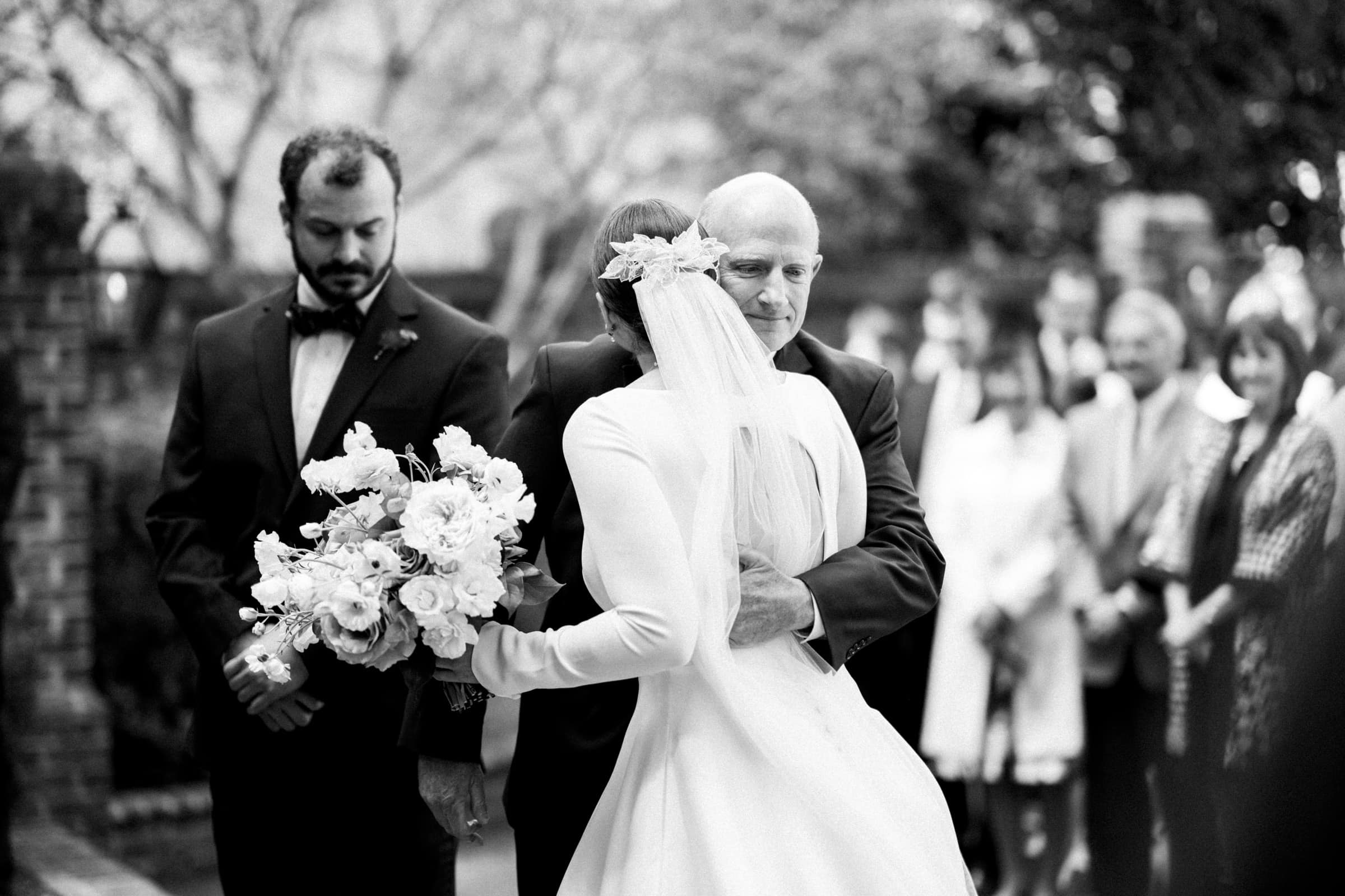 sutherland estate wedding - stephanie & hunter A black and white photo captures a bride in a gown and veil holding a bouquet as an older man, possibly her father, embraces her at the Sutherland Estate wedding. A groom in a suit stands to the side, with guests blurred in the background during the outdoor ceremony.