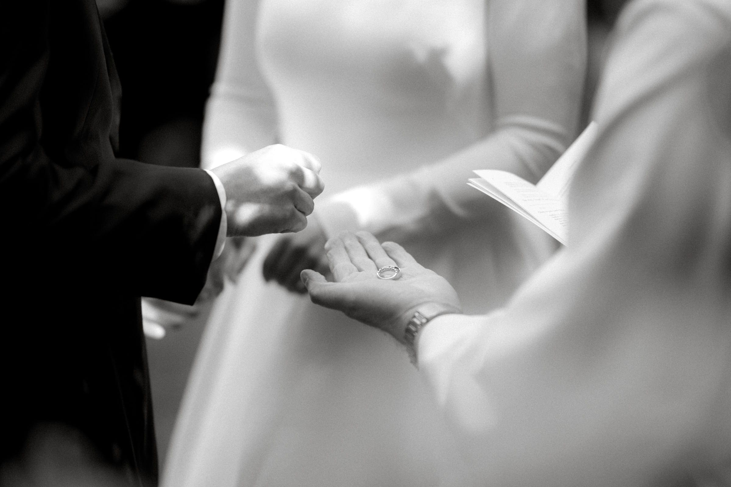 sutherland estate wedding - stephanie & hunter A black and white photo from a Sutherland Estate wedding captures a groom placing a ring on the bride's outstretched hand as vows are read from an open booklet. The bride, in her long-sleeved dress, stands in the softly lit ambiance of this charming venue.