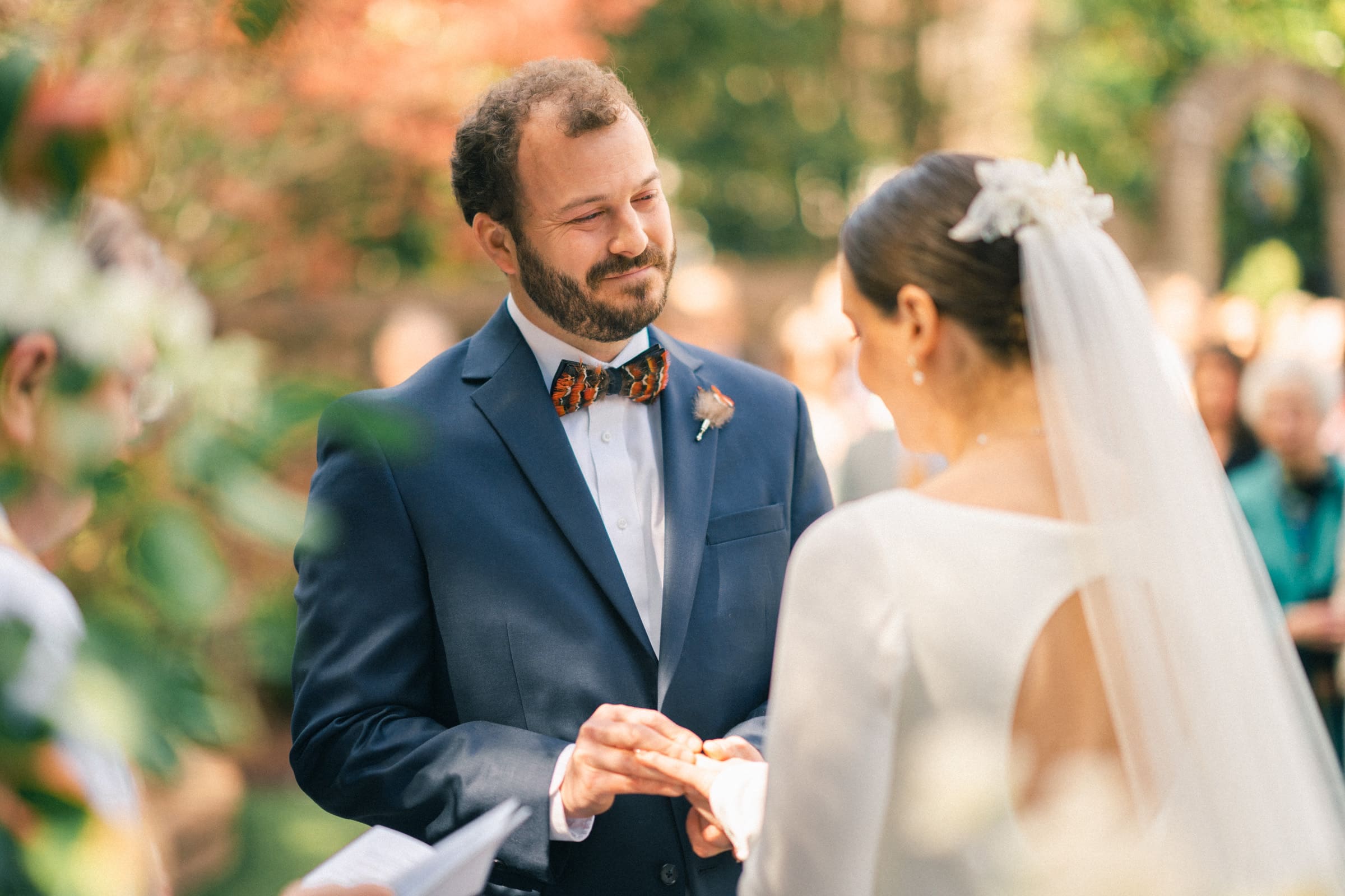 sutherland estate wedding - stephanie & hunter At a picturesque Sutherland Estate wedding, a groom in a navy suit and bow tie gently places a ring on the bride's finger during the outdoor ceremony. She looks stunning in her white dress and veil, surrounded by lush greenery and attentive guests.
