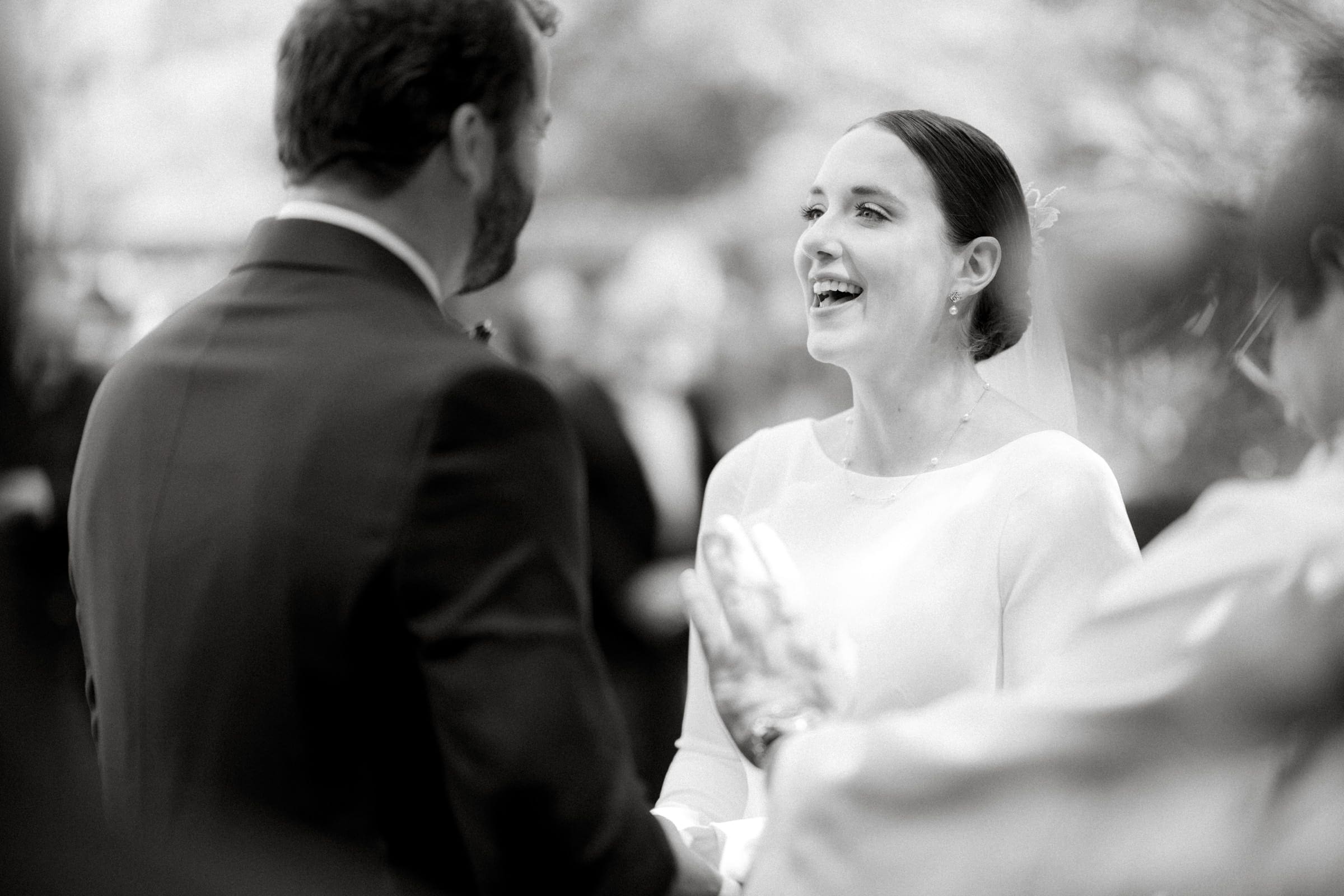 sutherland estate wedding - stephanie & hunter Black and white photo of a bride and groom smiling at each other during an outdoor Sutherland Estate wedding. The bride wears a veil and simple dress, while the groom is in a suit. Guests are blurred in the background.